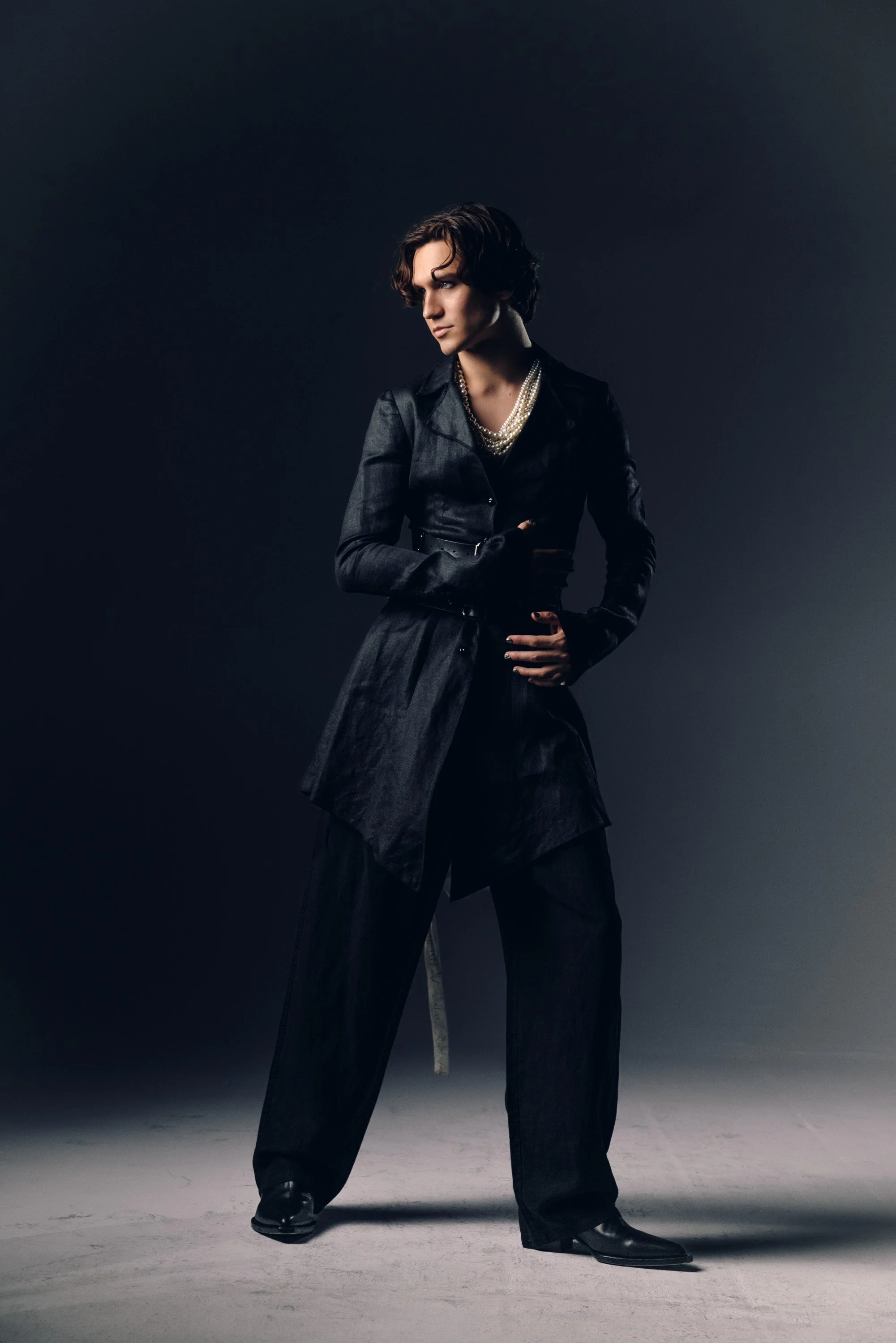 A woman with short curly hair in a dark setting wearing a black trench coat, layered necklaces, black pants, and black boots.