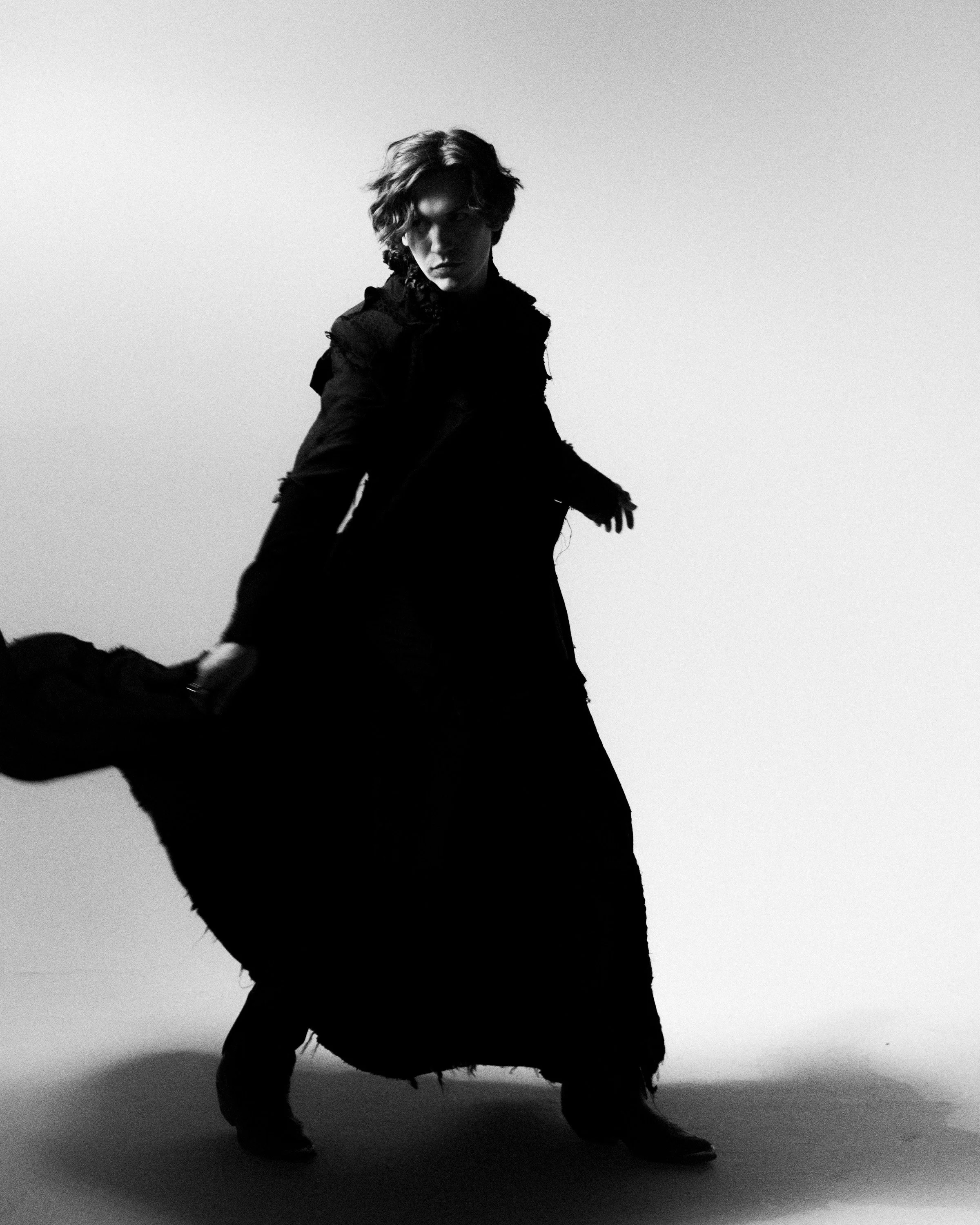 A black-and-white photo of a person with medium-length, tousled hair, wearing dark clothing, standing in front of a plain background. The person appears to be in a dynamic pose with one arm extended and a serious expression.