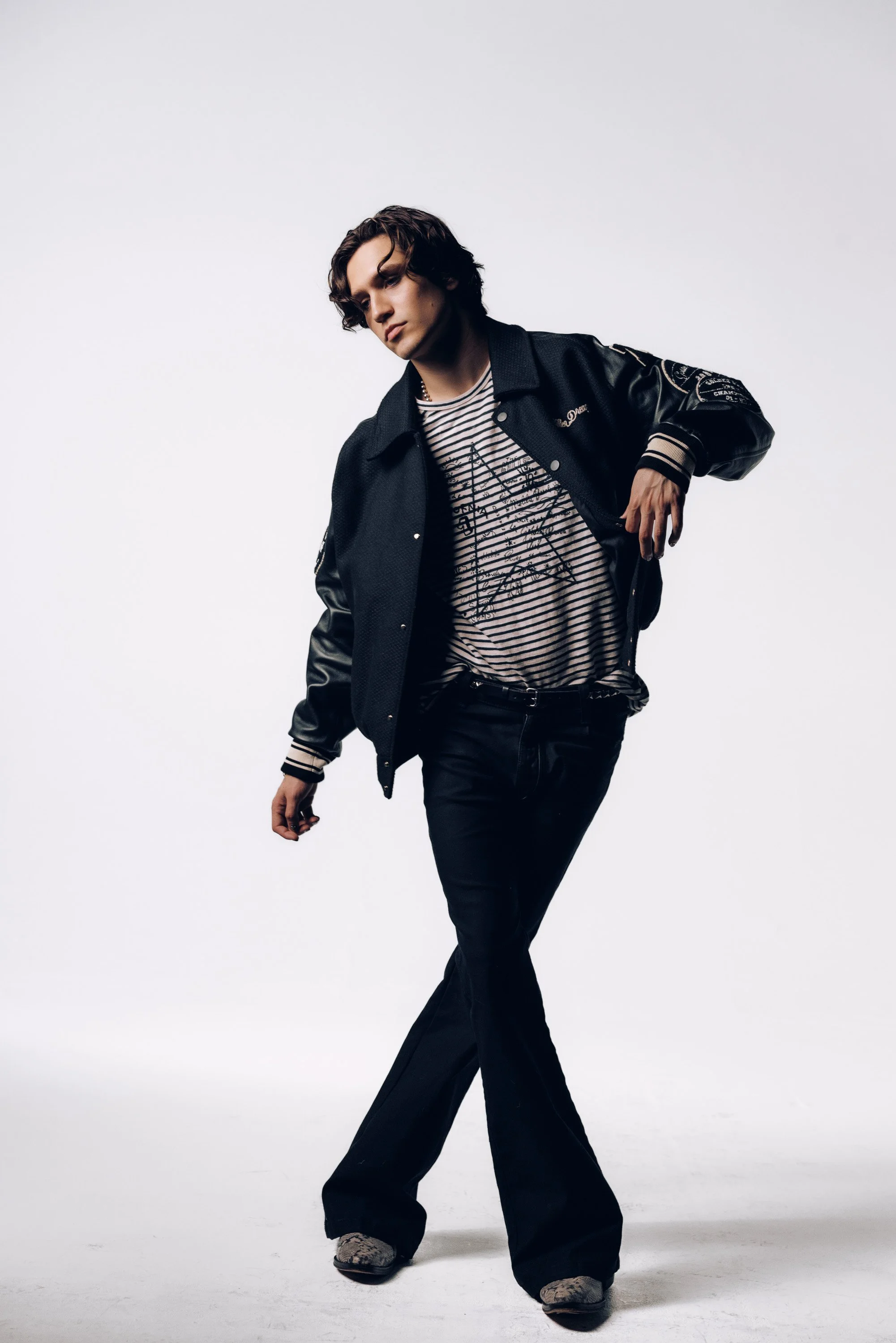 A young man with wavy brown hair wearing a black varsity jacket with leather sleeves, a striped T-shirt, black pants, and sneakers standing against a plain white background, facing slightly to the left and looking downward.