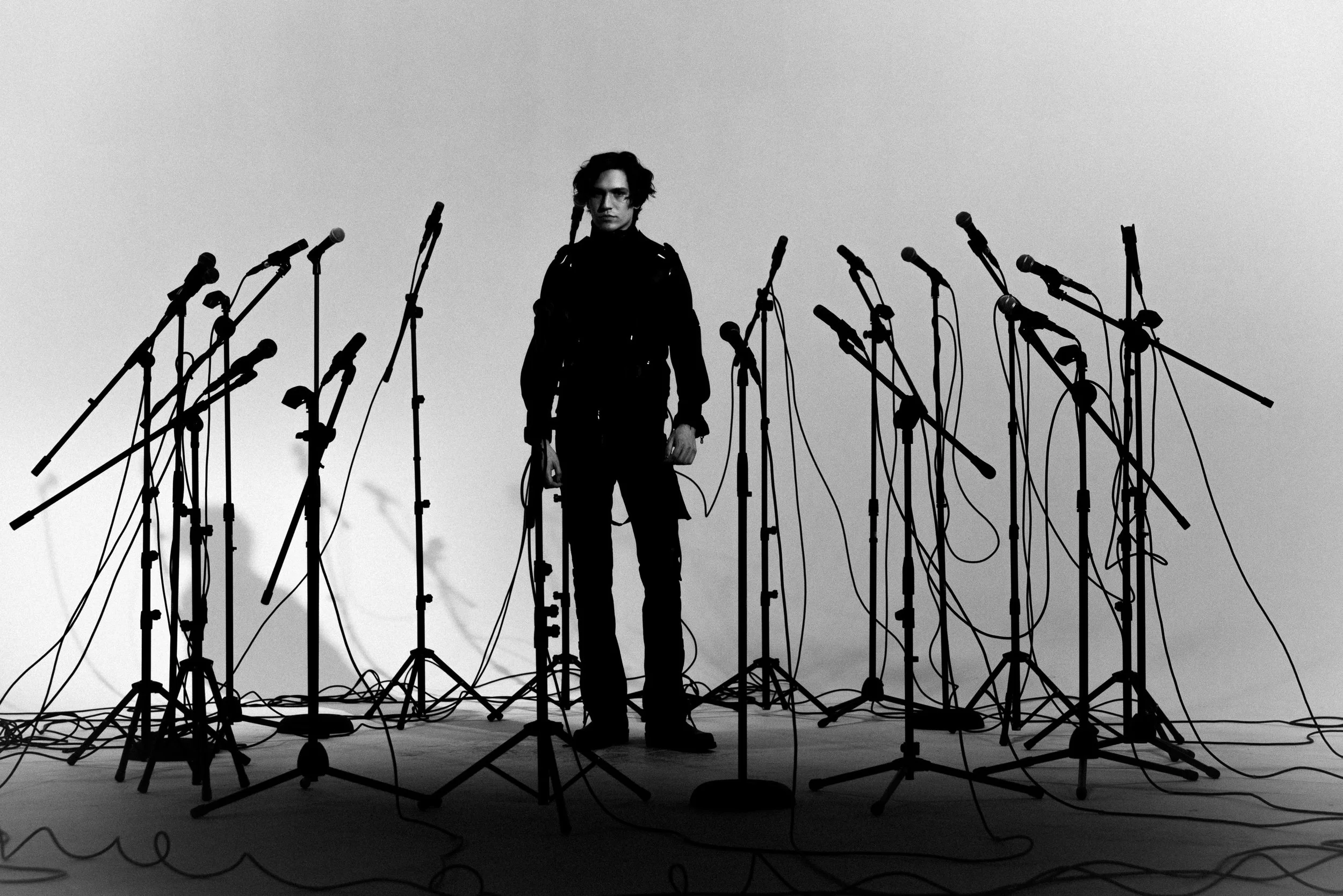 A person standing among numerous microphones on stands, set on a stage or studio, against a plain, light background.