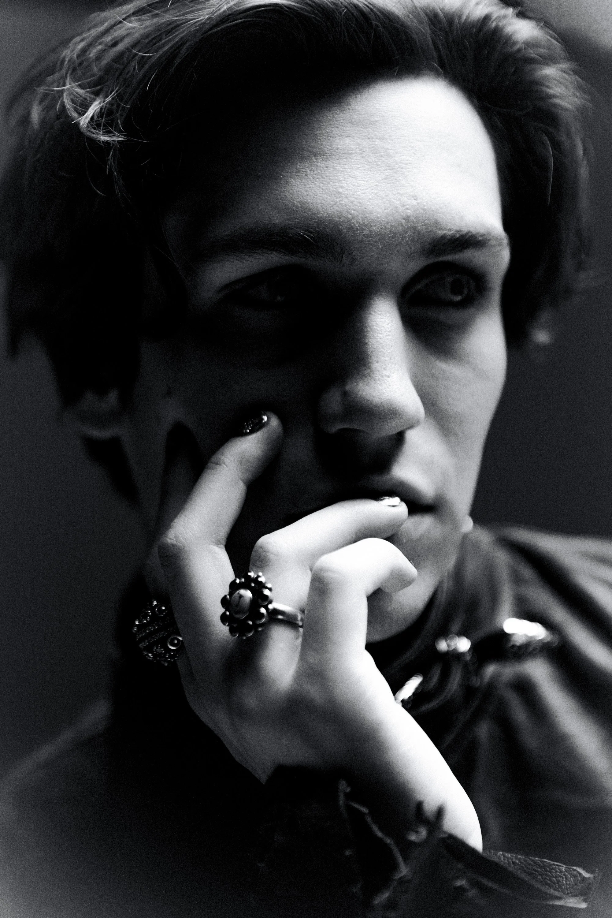 Close-up of a young person's face in black and white, with focus on one eye and hand near the face, wearing rings and bracelets.