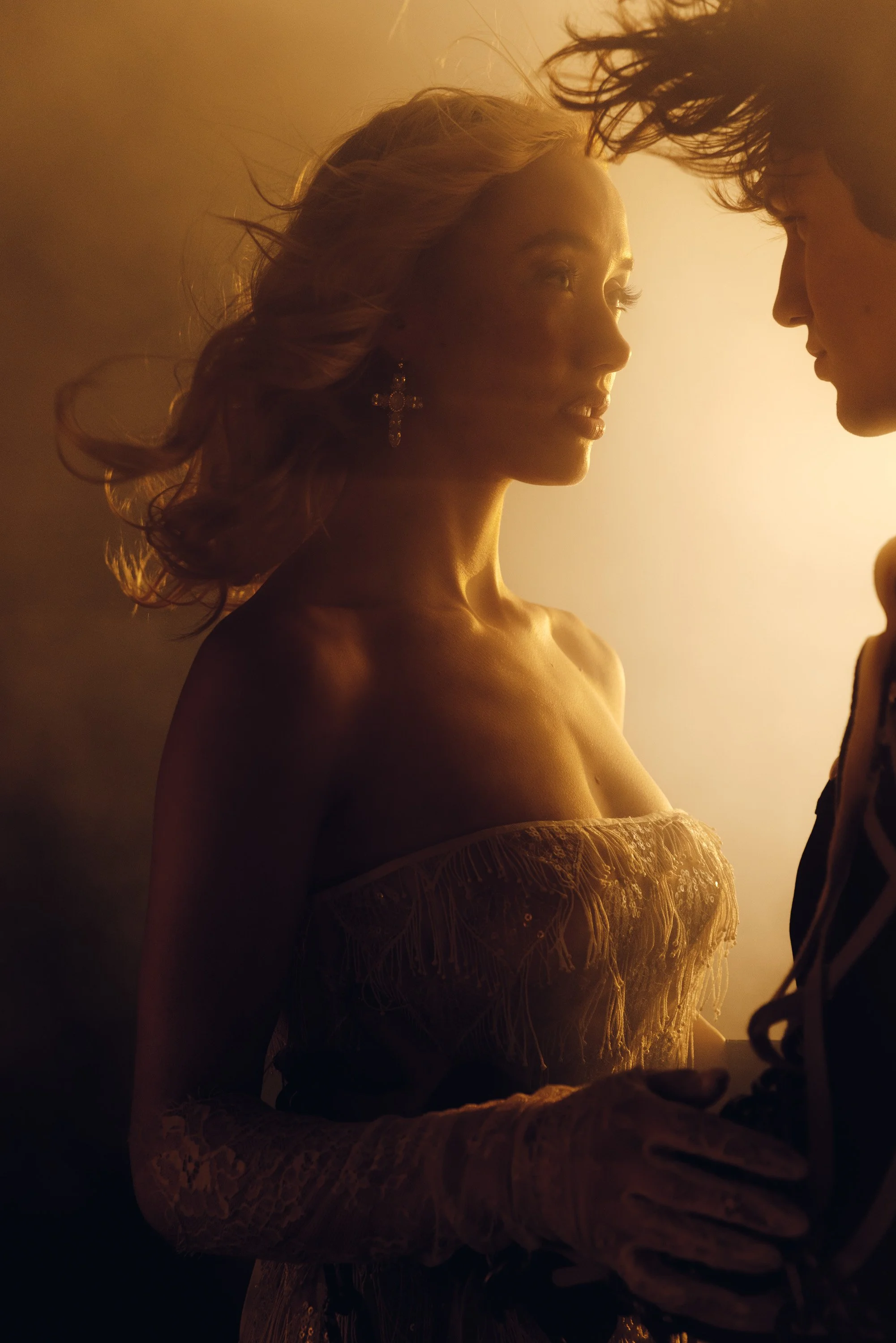 A woman with long, wavy hair and earrings, and a person with curly hair and glasses, face each other close in warm, soft lighting.