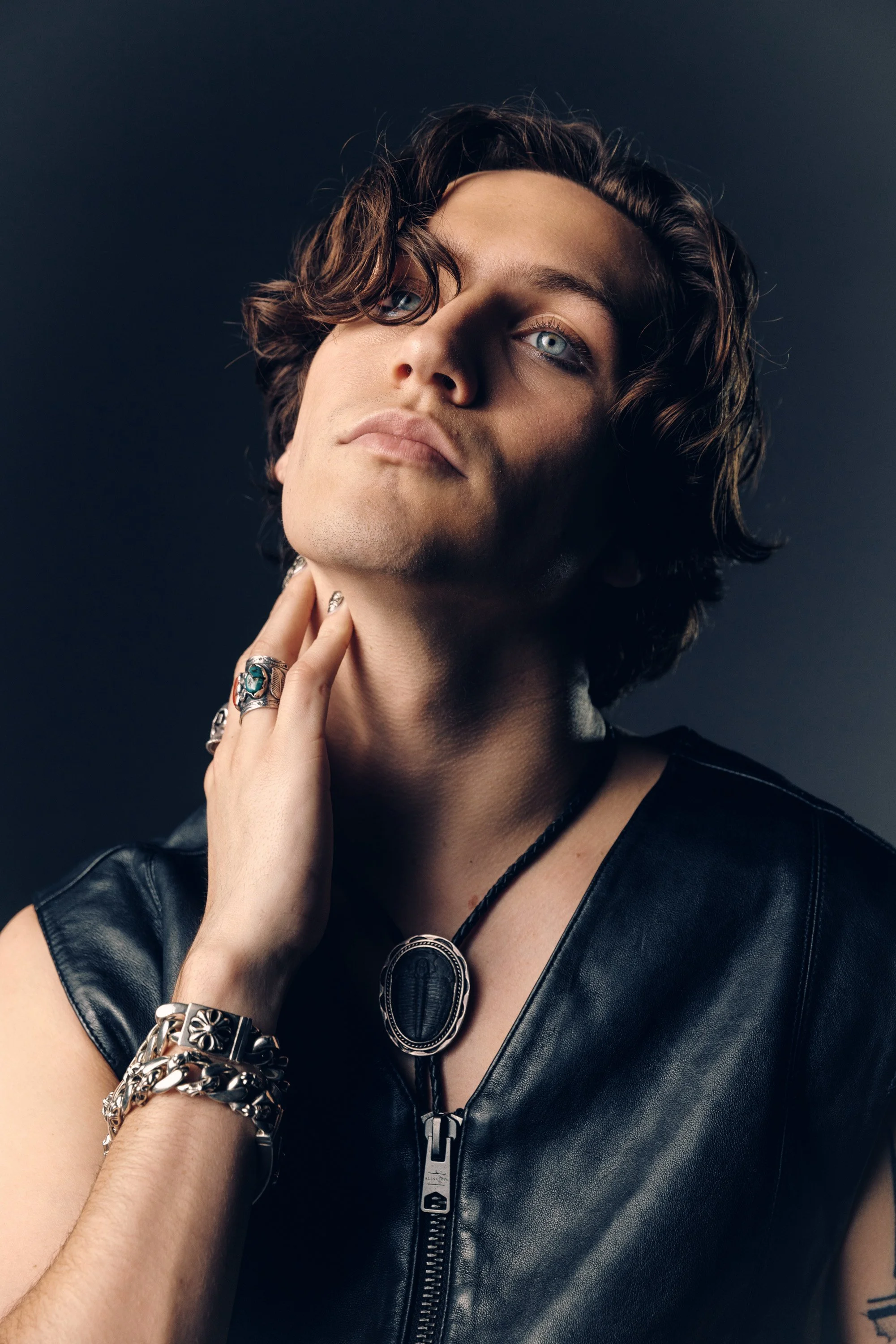A person with blue eyes and curly brown hair touching their neck, wearing a black leather vest, multiple rings, bracelets, and a necklace with a large circular pendant, against a dark background.
