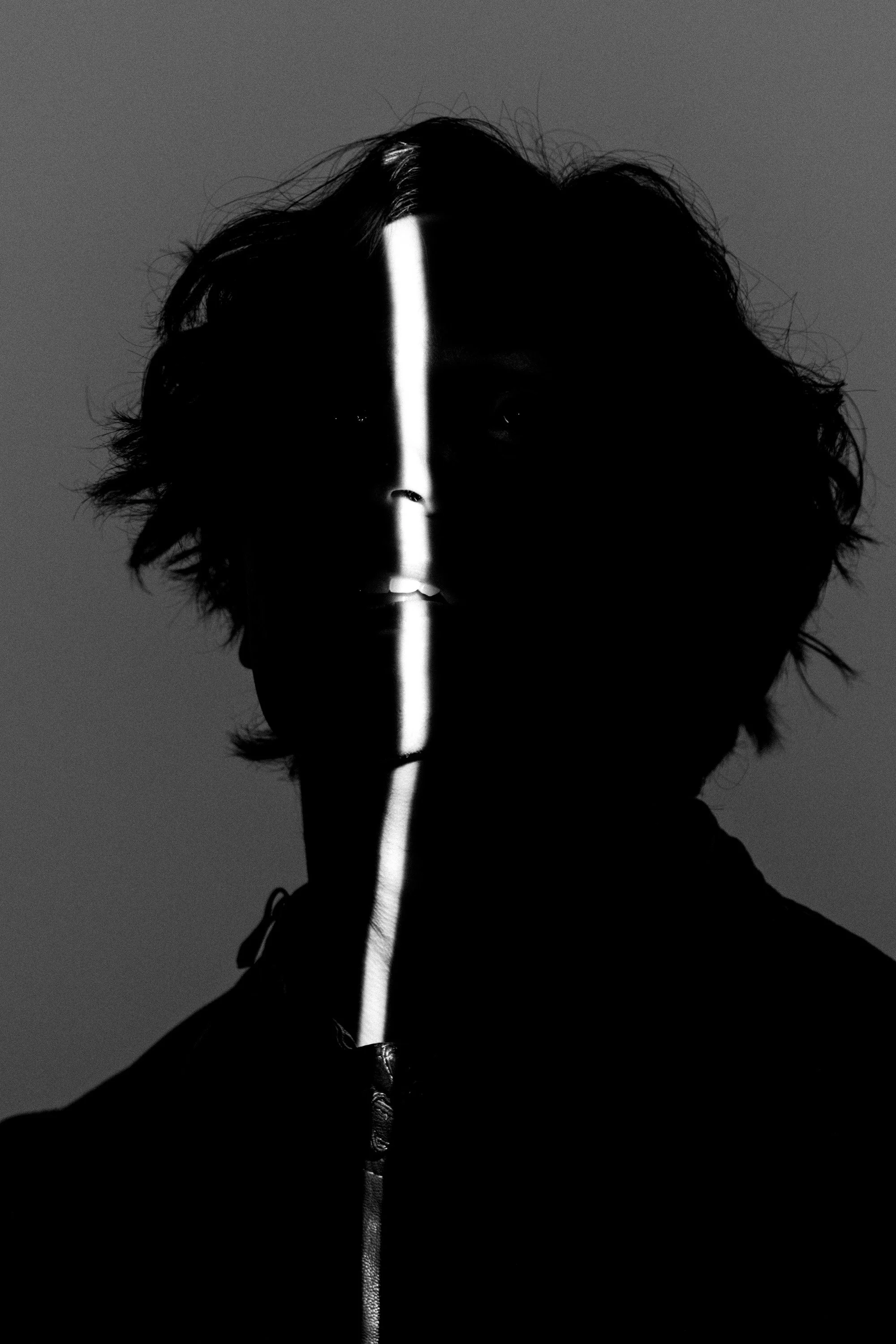 Black and white photo of a person with curly hair, facing forward, with a bright vertical line of light on their face and chest, creating a high-contrast, abstract effect.