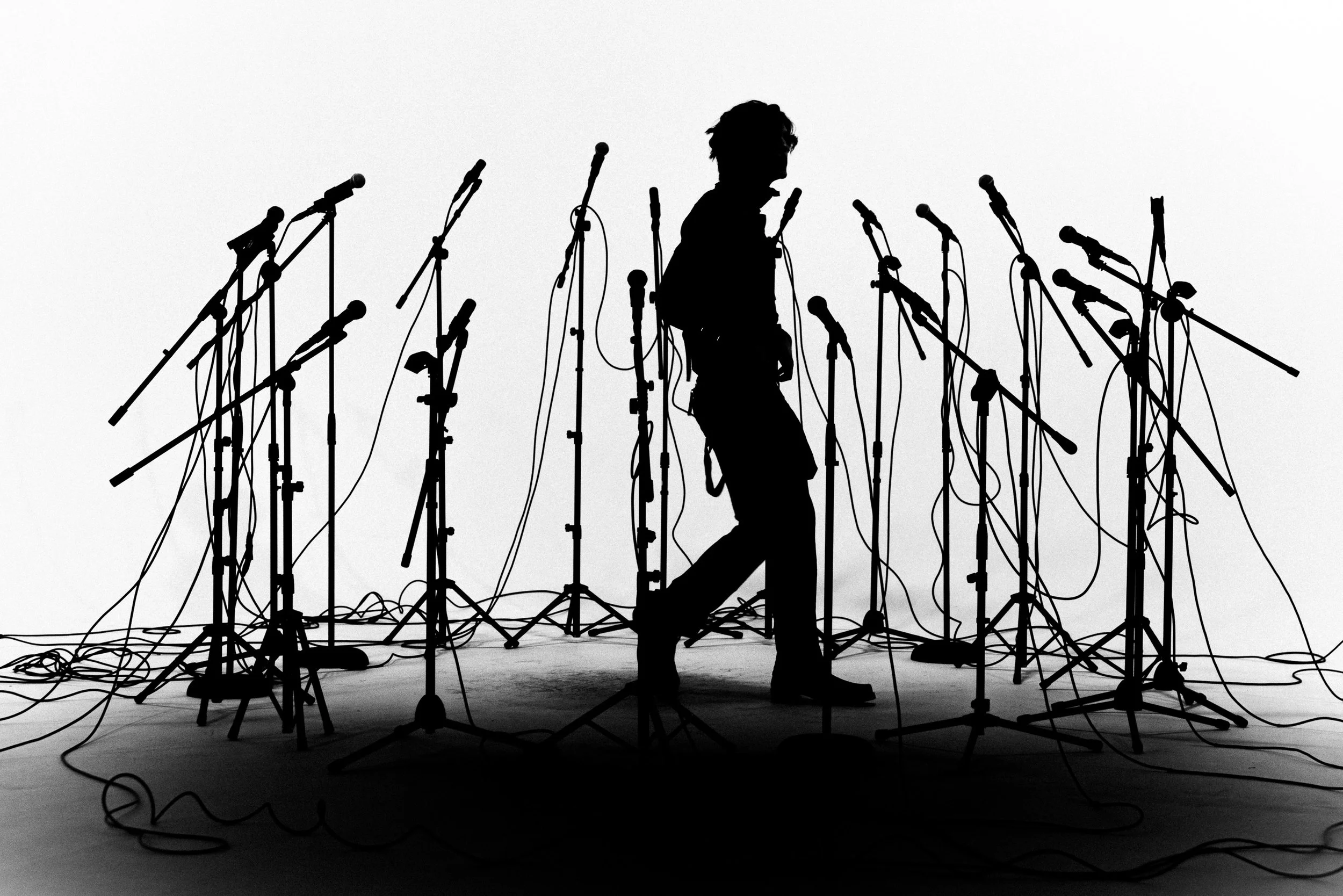 Silhouette of a person walking among numerous microphone stands on a stage with tangled wires.