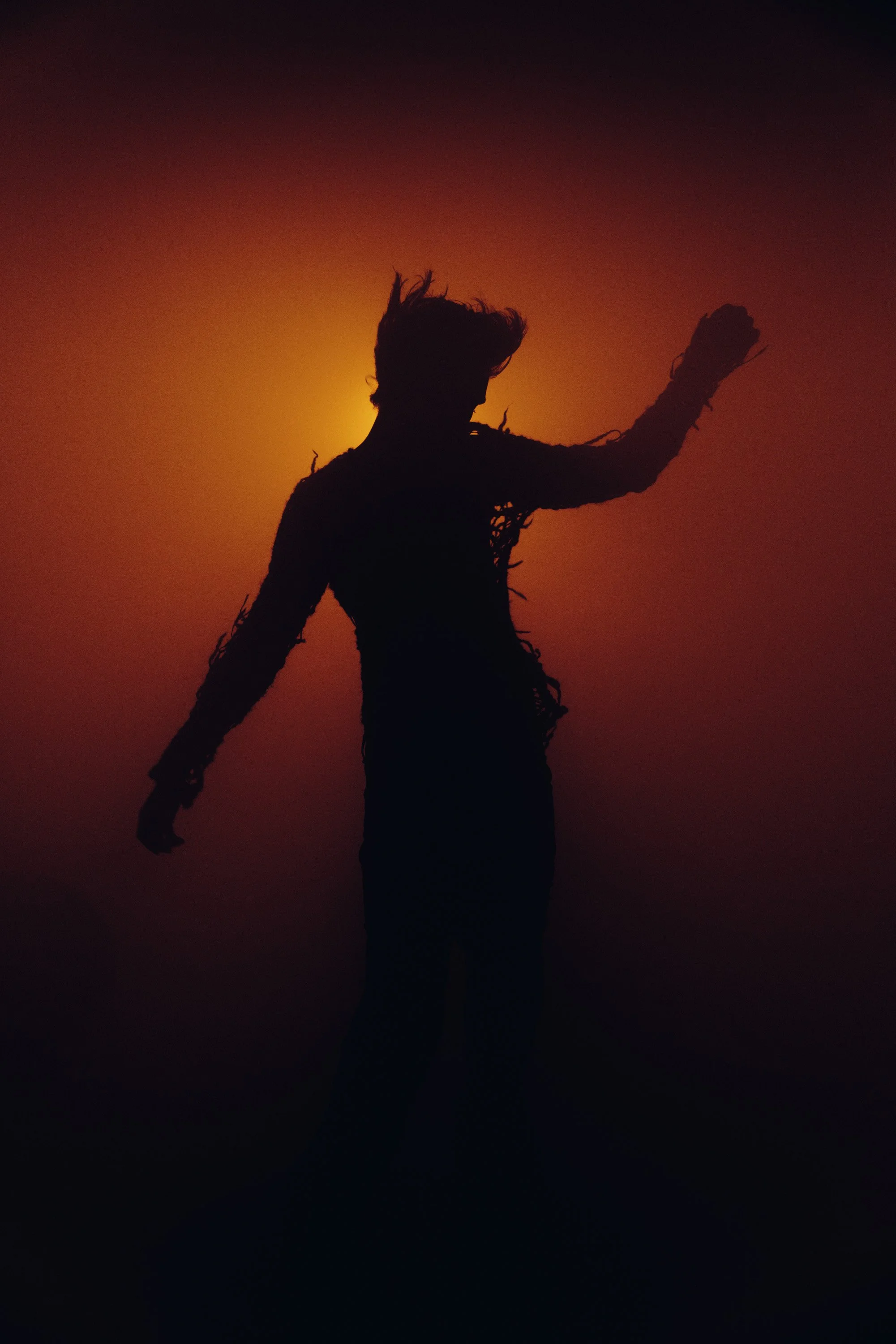 Silhouette of a person waving with arm raised against a sunset or sunrise background with a gradient of dark orange, red, and purple hues.