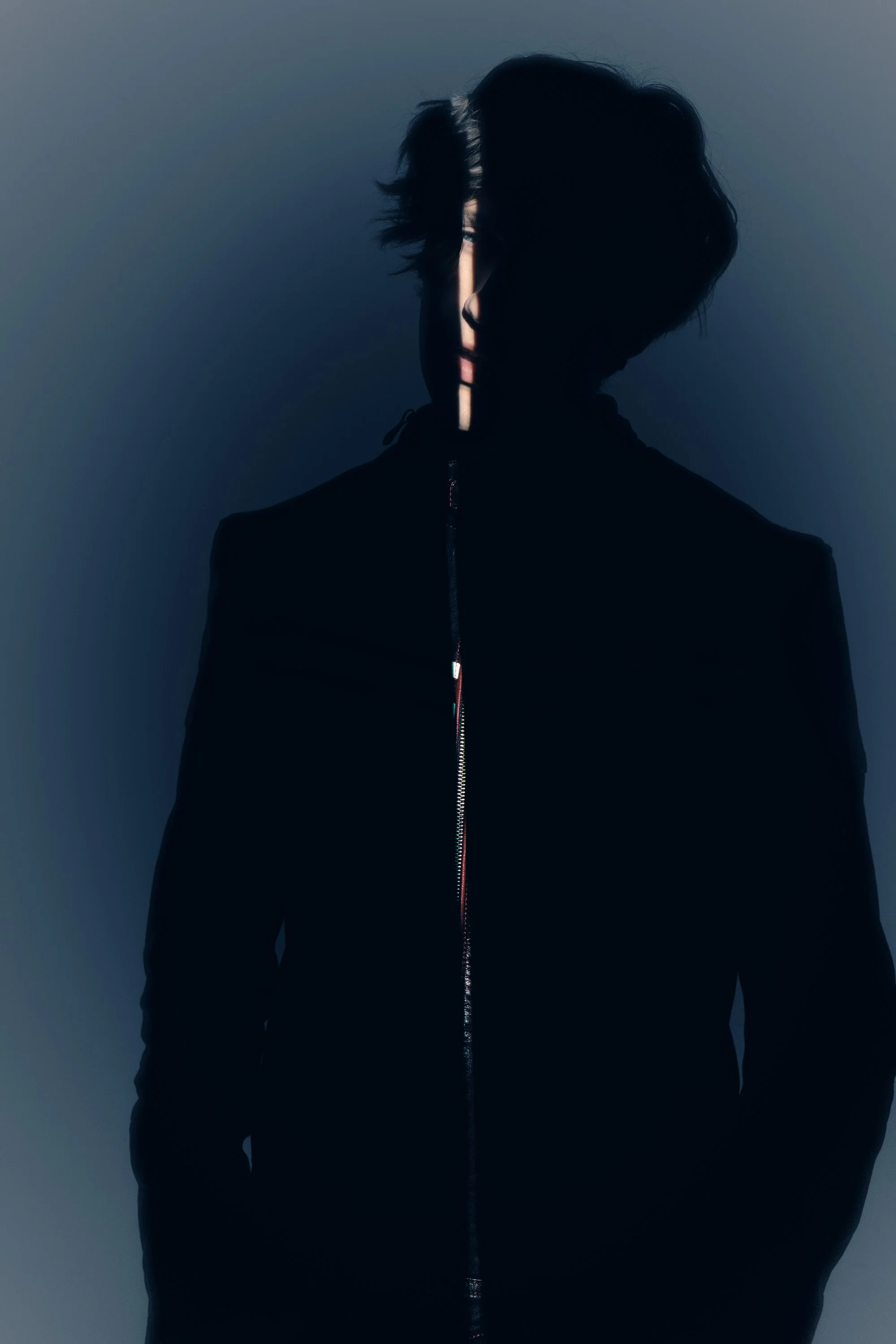 Silhouette of a person with wavy hair, wearing a black jacket, facing forward with face partially obscured by light and shadow.