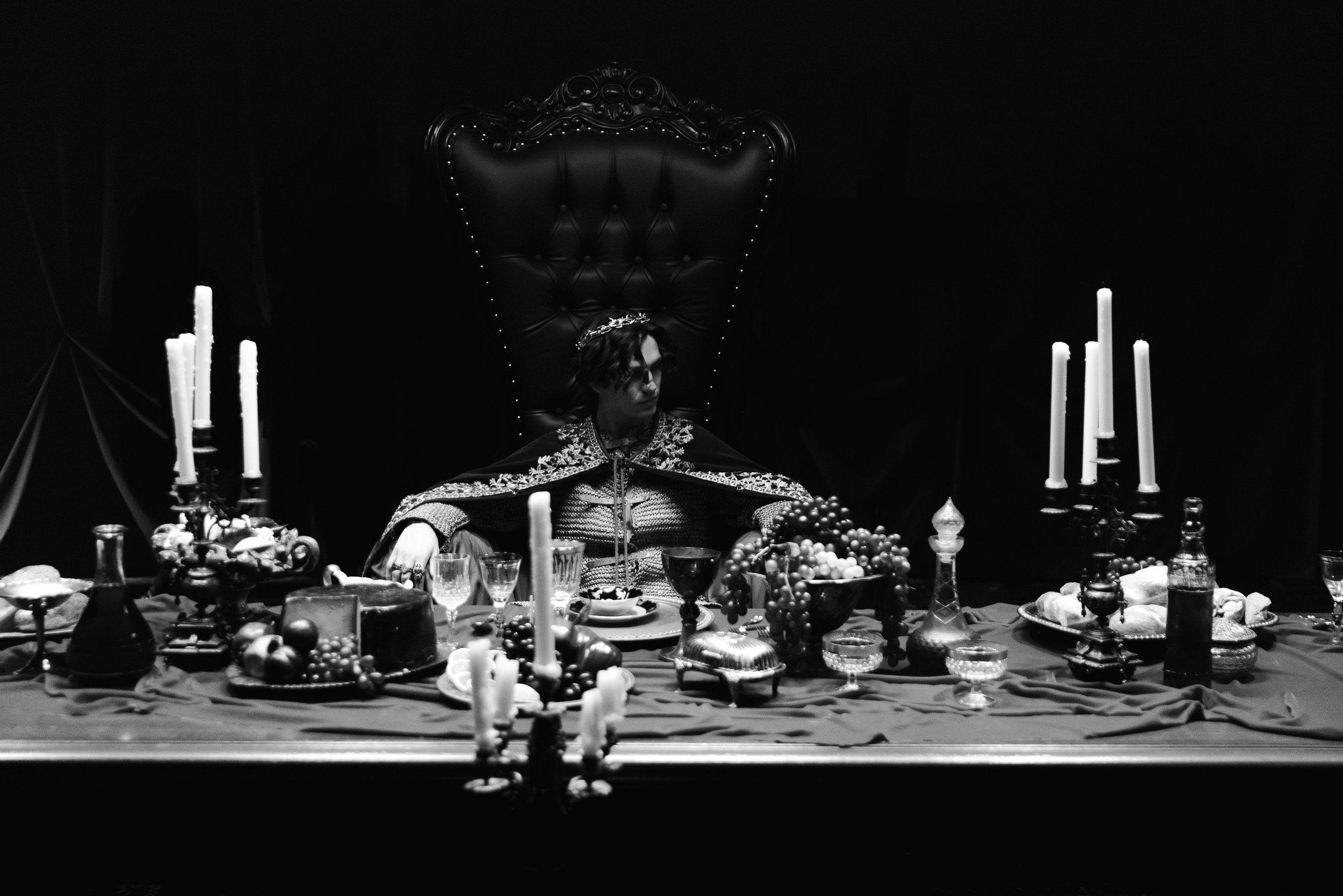 A person dressed in ceremonial clothing, wearing a crown, sitting at an elaborately decorated table with candles and various food items, against a dark background with a high-backed ornate chair.