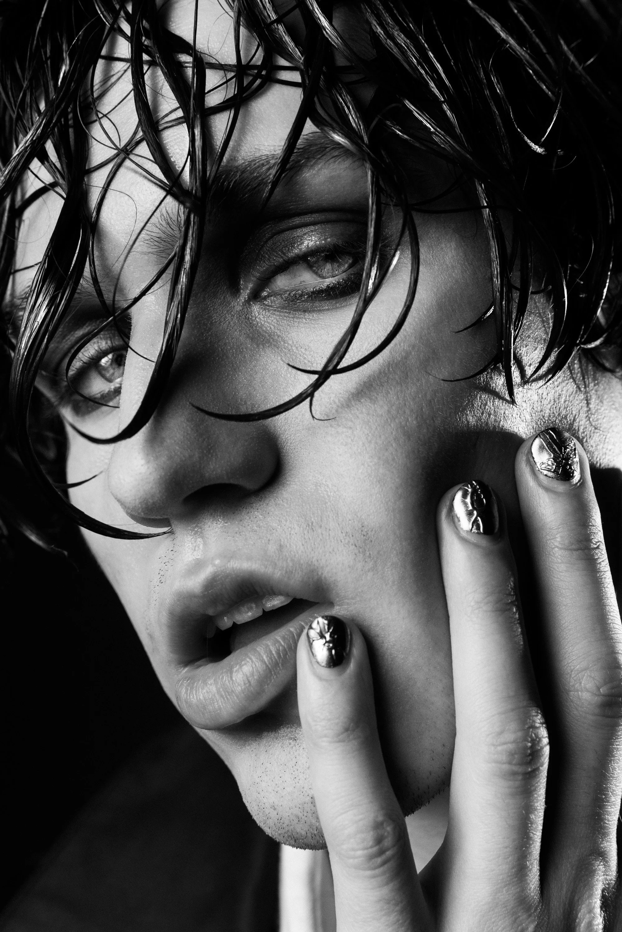 Close-up black and white photo of a woman with wet hair partially covering her face, making an intense expression with her lips slightly parted and fingers touching her lips.
