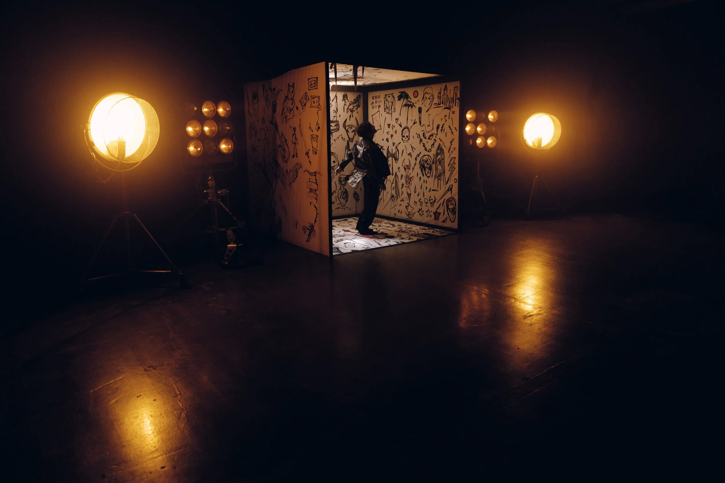 A person walking inside a small booth decorated with black doodle-style drawings on white walls, illuminated by two large studio lights on either side.