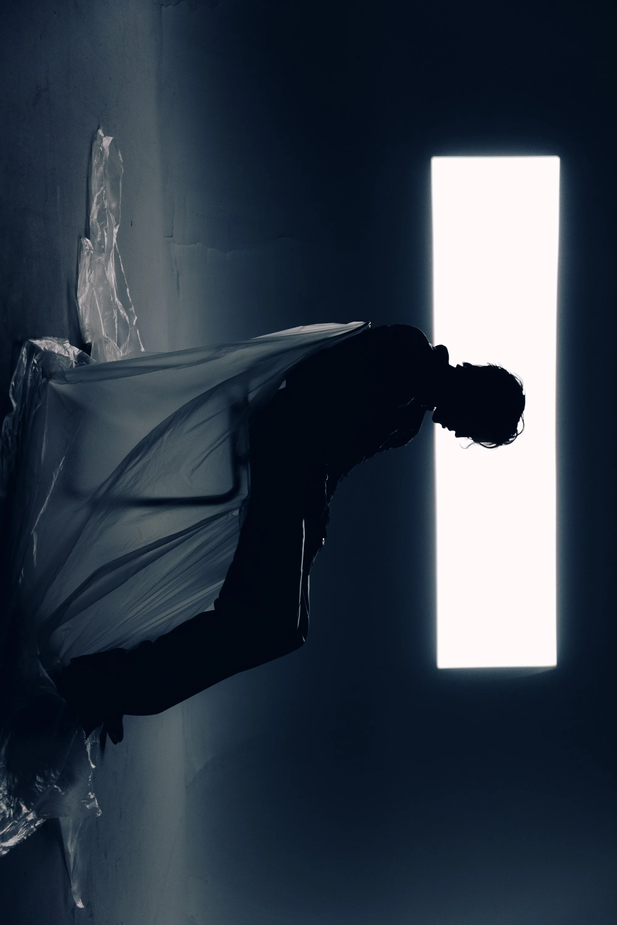 A person standing in front of a bright rectangular window in a dark room, with plastic sheets on the floor.