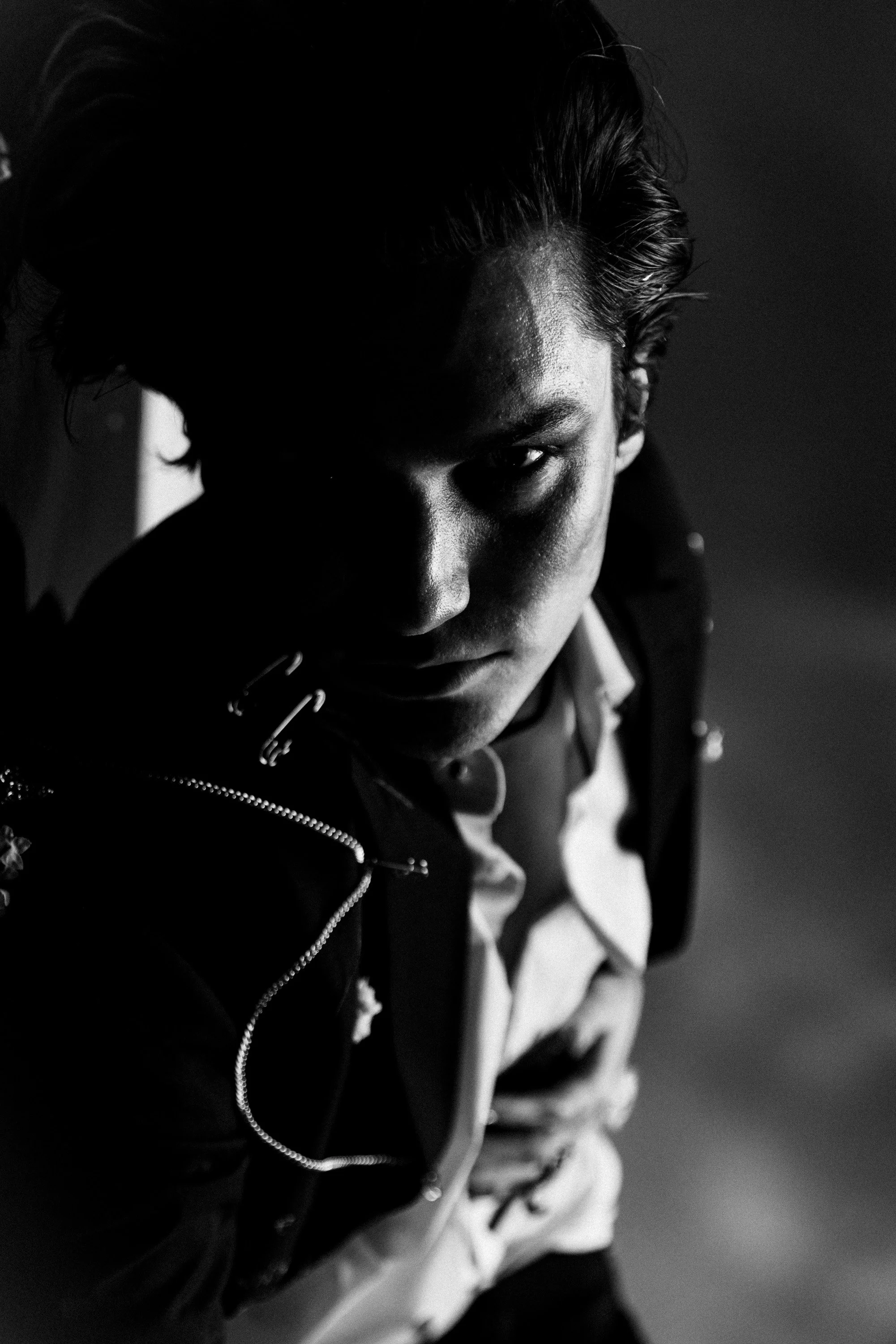 Black and white close-up portrait of a young man with styled hair, wearing a zip-up jacket and a shirt, looking intensely at the camera.