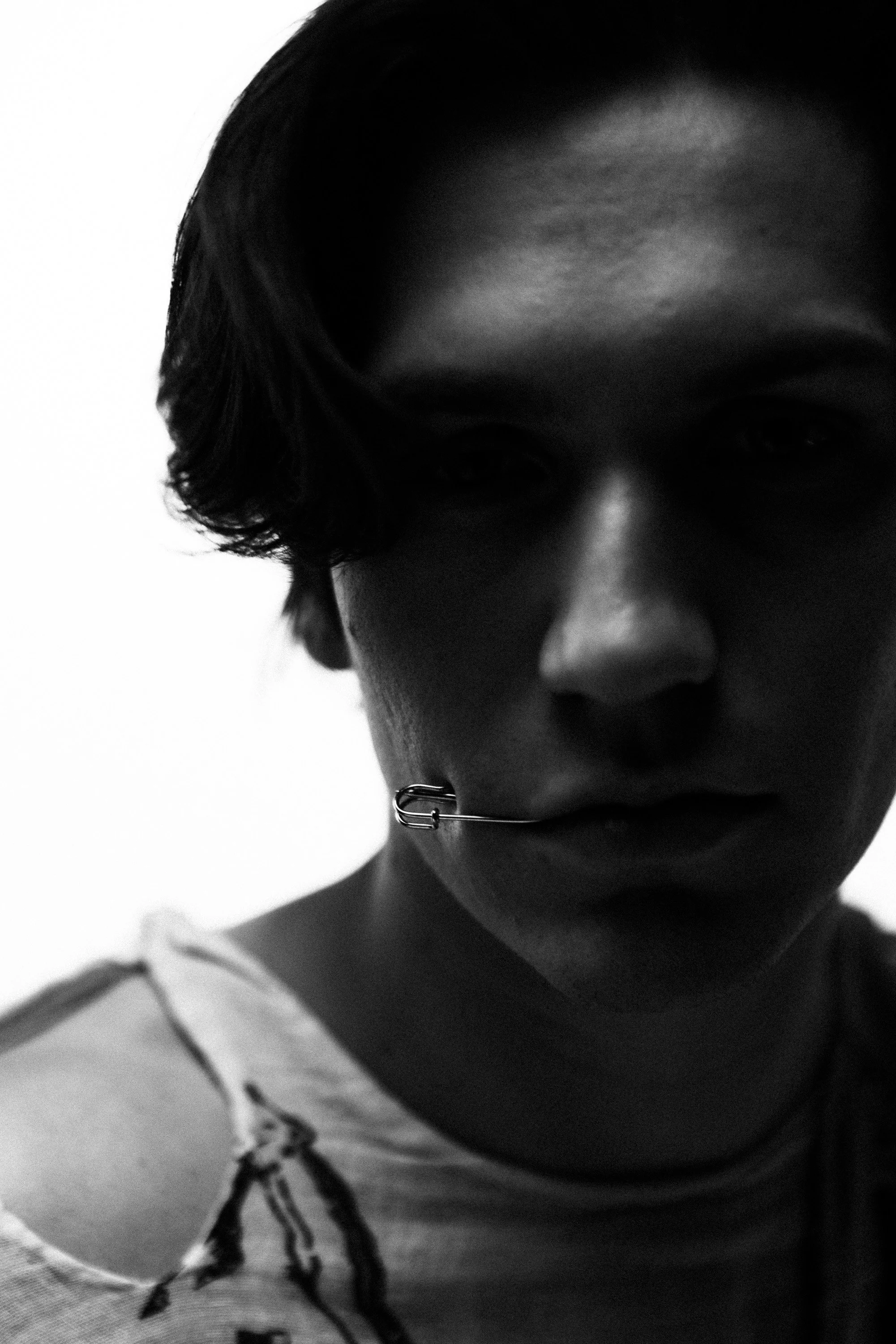 Close-up black and white photo of a young person's face, showing part of their hair, eye, and nose, with a piece of jewelry in their nose, and their clothing visible at the bottom of the image.