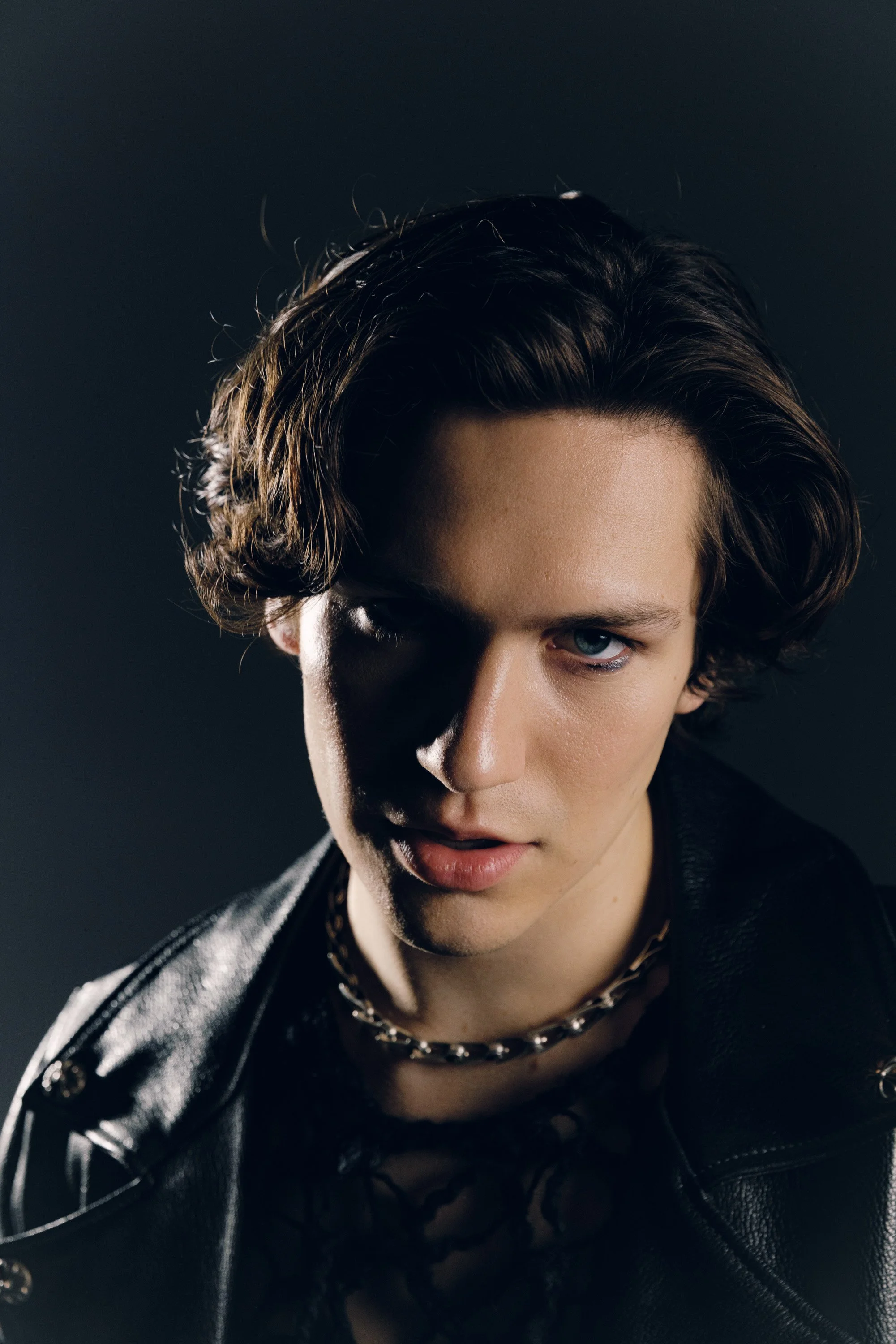 Close-up of a person with dark, wavy hair and piercing blue eyes, wearing a black leather jacket and a layered chain necklace, with a dark background.