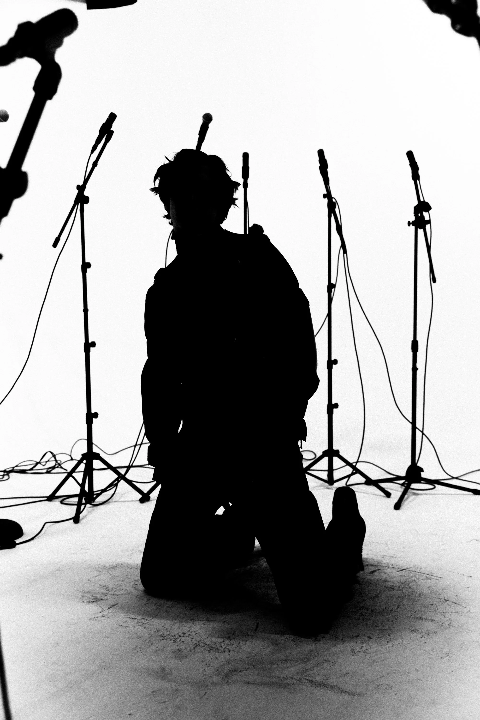Silhouette of a person kneeling on the floor in front of multiple microphones on stands, with cables on the ground, against a white background.