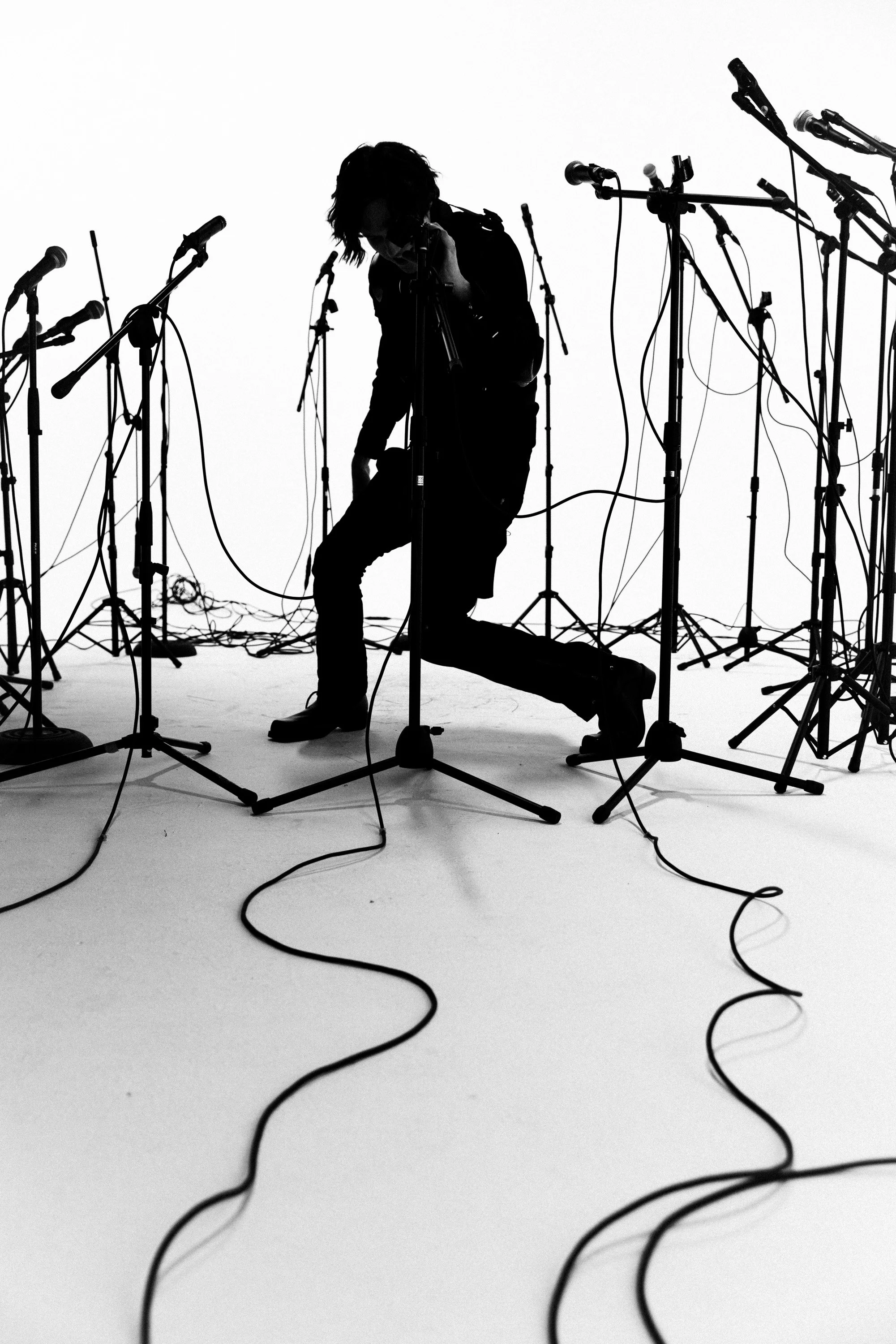 Silhouette of a person kneeling and leaning forward among numerous microphones on stands, with many cables on the ground, all against a white background.
