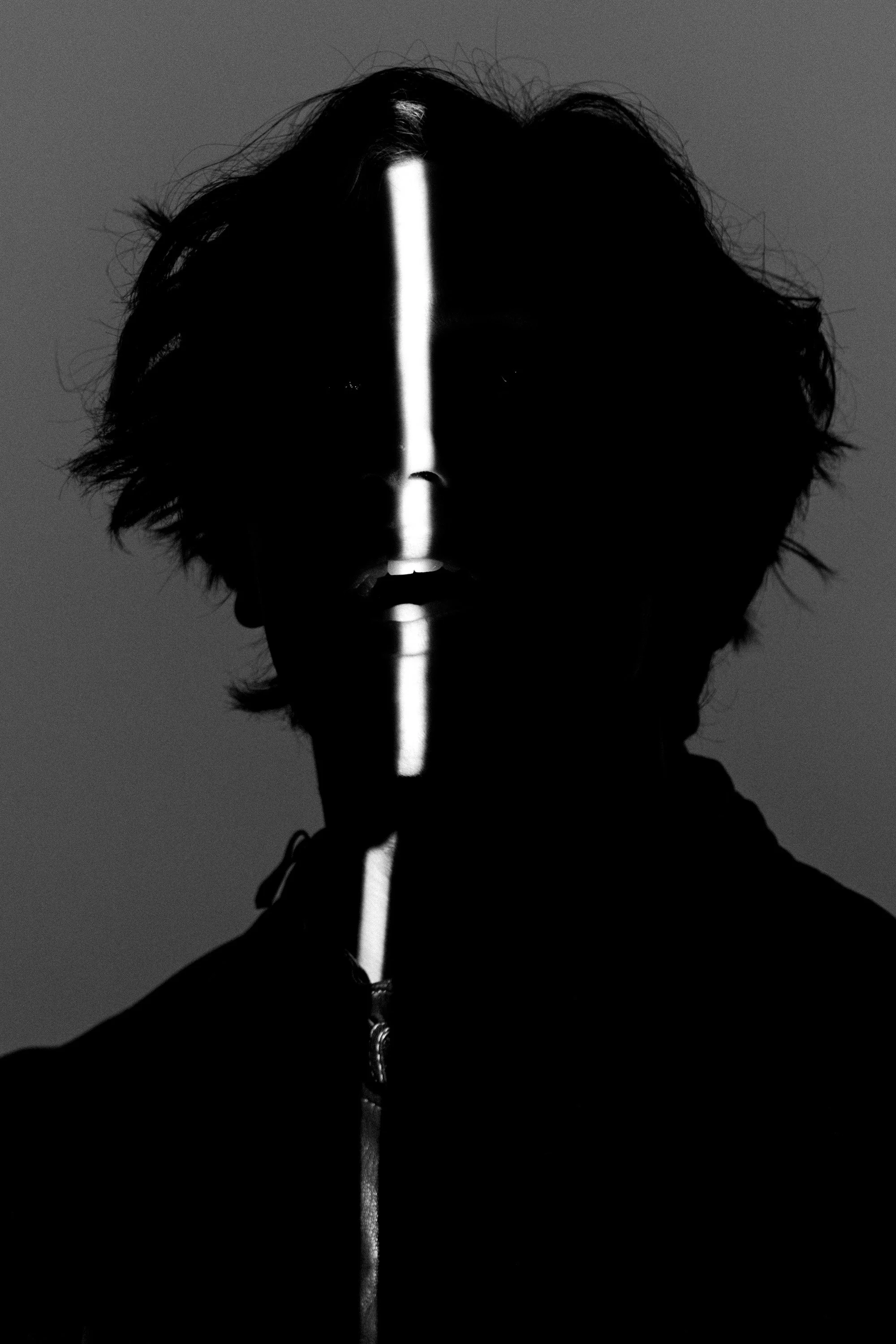 Black and white silhouette of a person with messy hair, facing forward, with a vertical bright light streak down the middle of their face and body, set against a plain background.