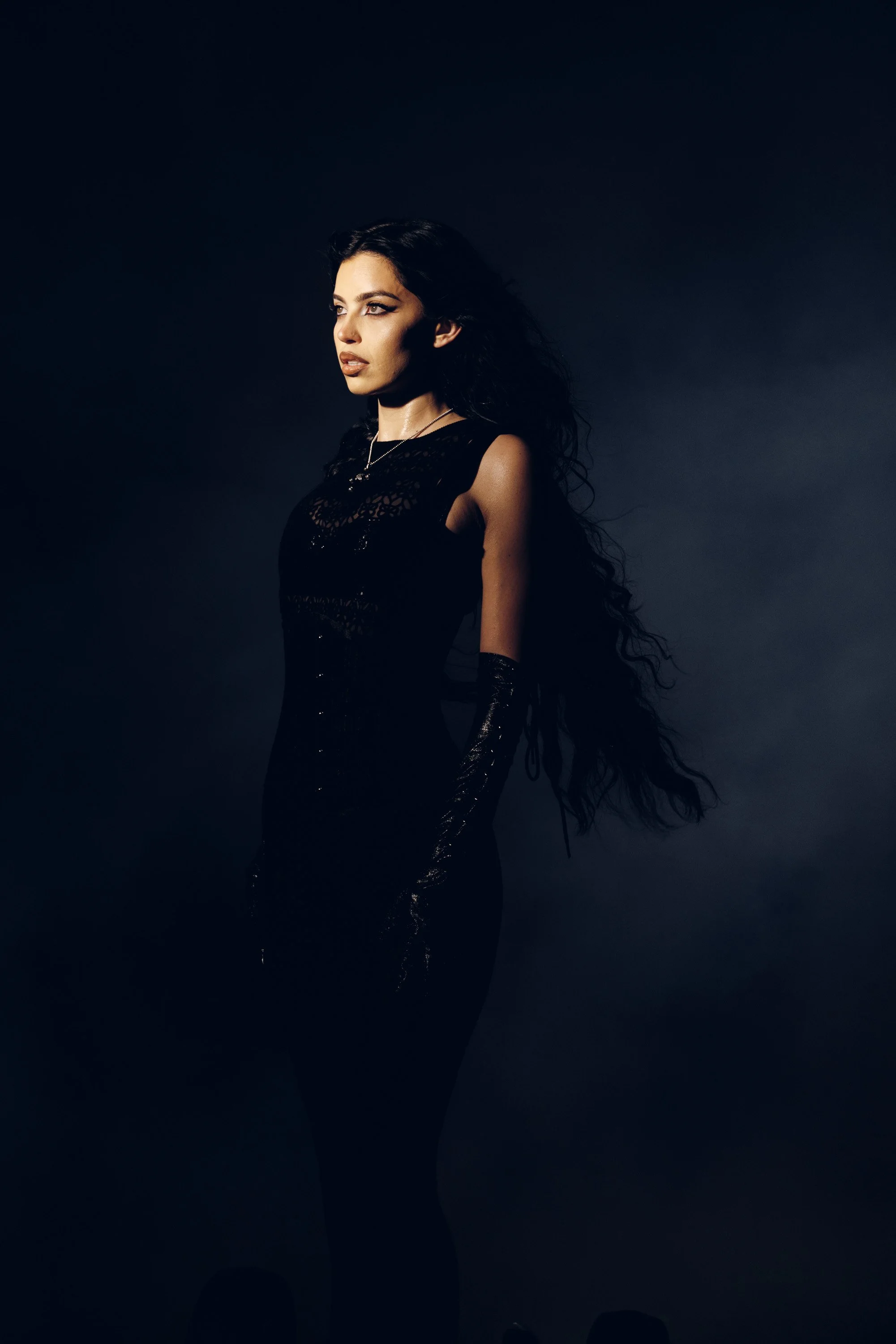 A woman with long black curly hair and medium skin tone looking to the side against a dark background, wearing a black dress with lace detail and gloves.