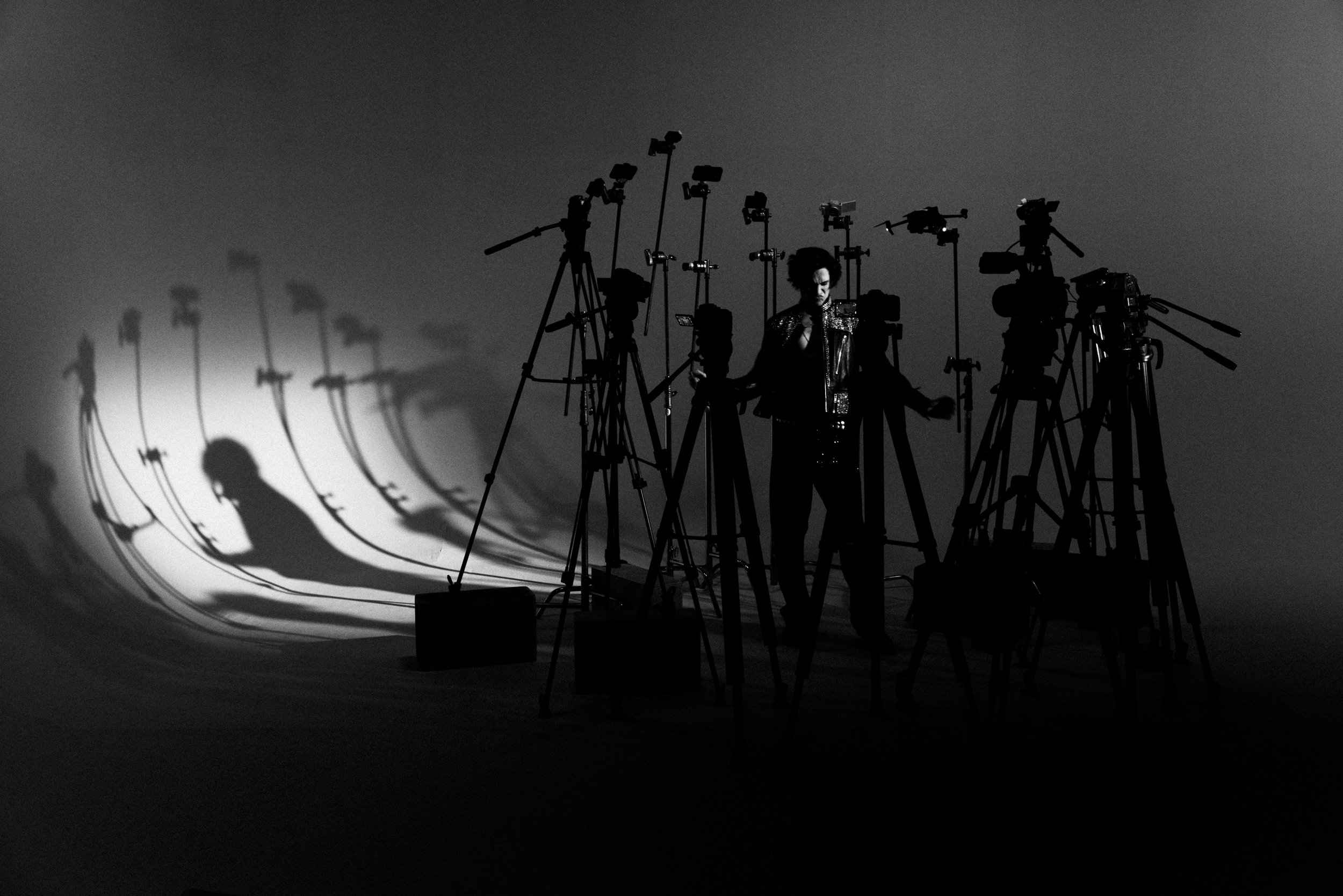 A person with curly hair is surrounded by multiple cameras and tripods, with shadows cast on the wall behind them in a photography studio setting.