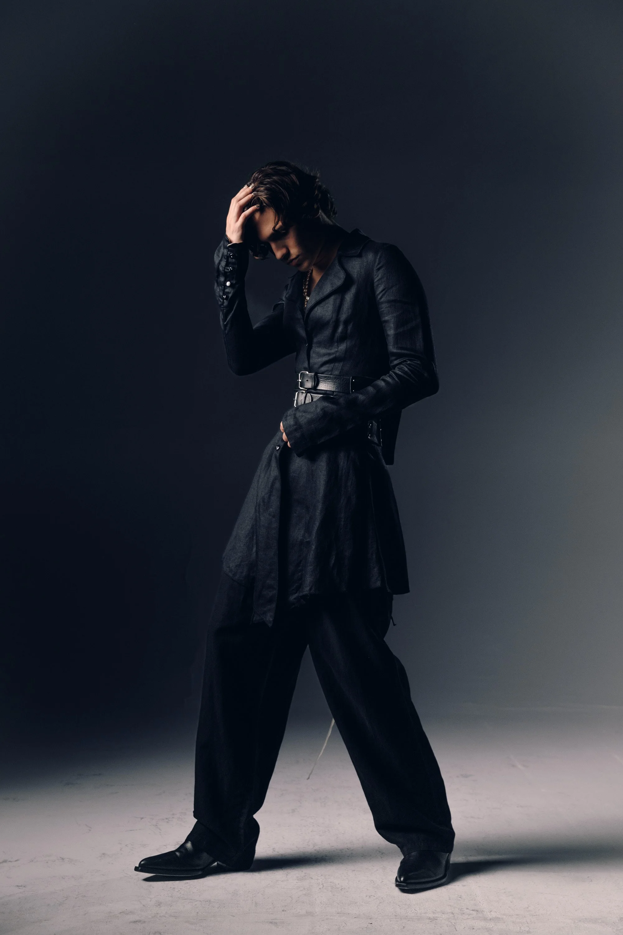 A person dressed in black standing against a dark background with one hand on their head, wearing a leather jacket, loose pants, and pointed shoes, striking a pose.