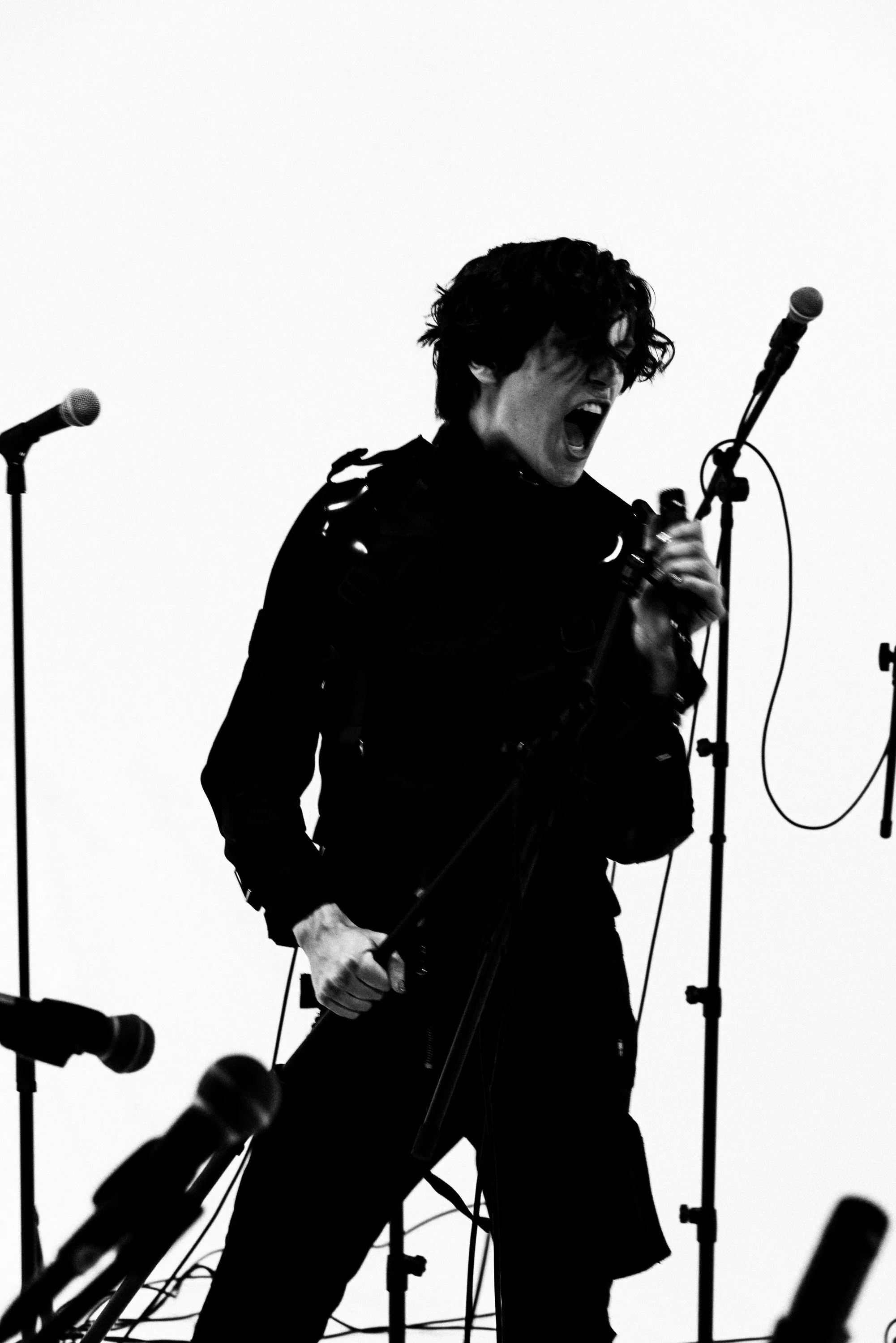 Black and white photograph of a person with curly hair passionately singing into a microphone, surrounded by multiple microphone stands and cables.