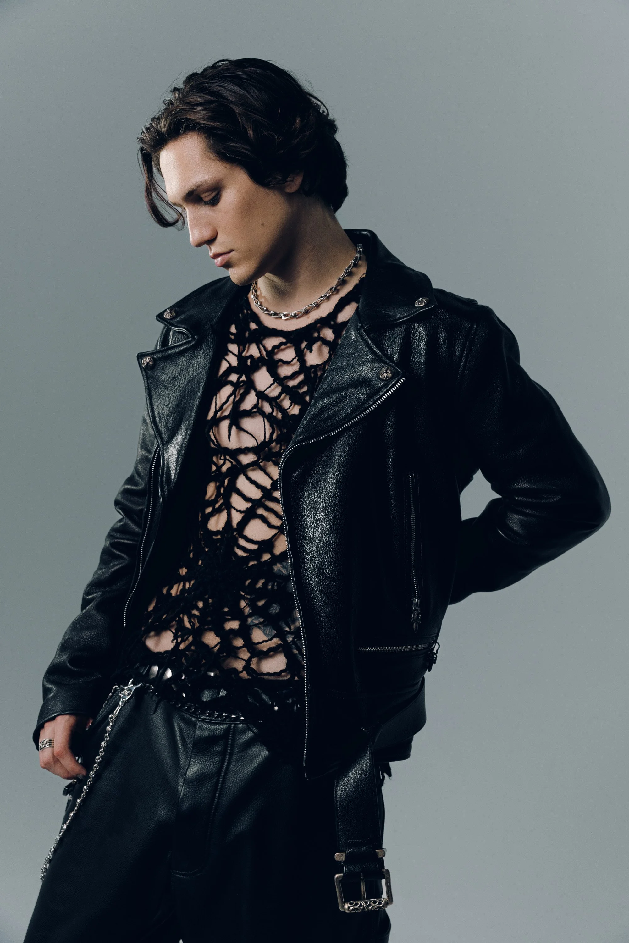 A person with short dark hair wearing a black leather jacket, a netted black top, and a silver chain necklace, standing against a plain gray background.
