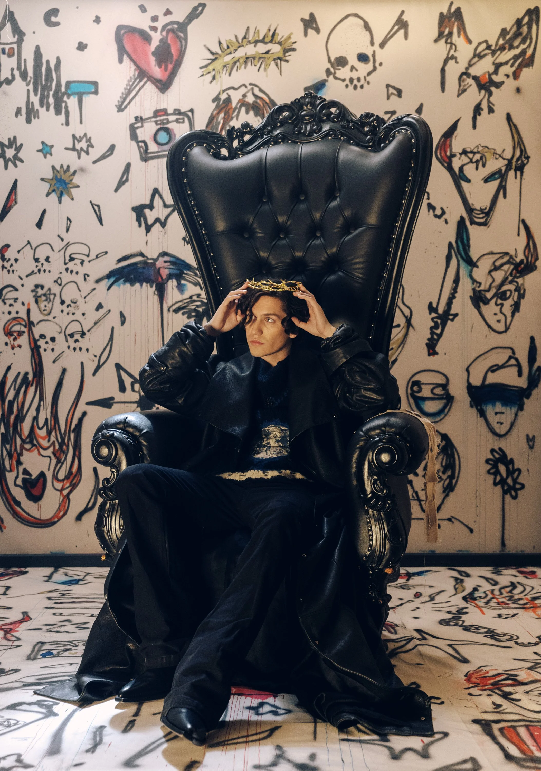 A person wearing a black leather jacket and pants sitting on a black ornate throne-like chair, holding a golden crown on their head. The background features a graffiti-style mural with various sketches, including a heart with an arrow, a skull, a cam