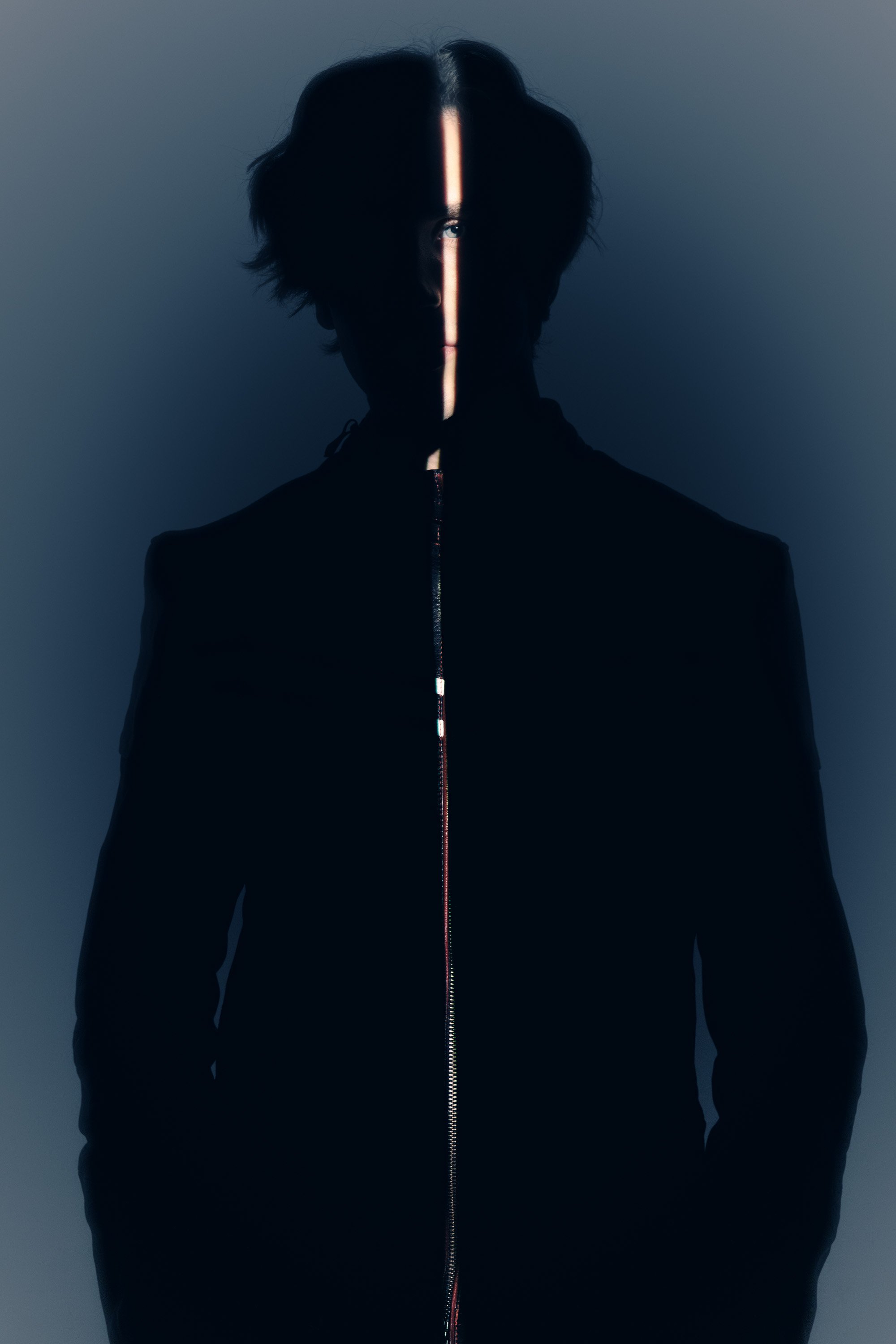 A person with black hair and light-colored eyes, dressed in dark clothing, stands against a plain background. A vertical light source divides the face, creating a dramatic shadow effect.