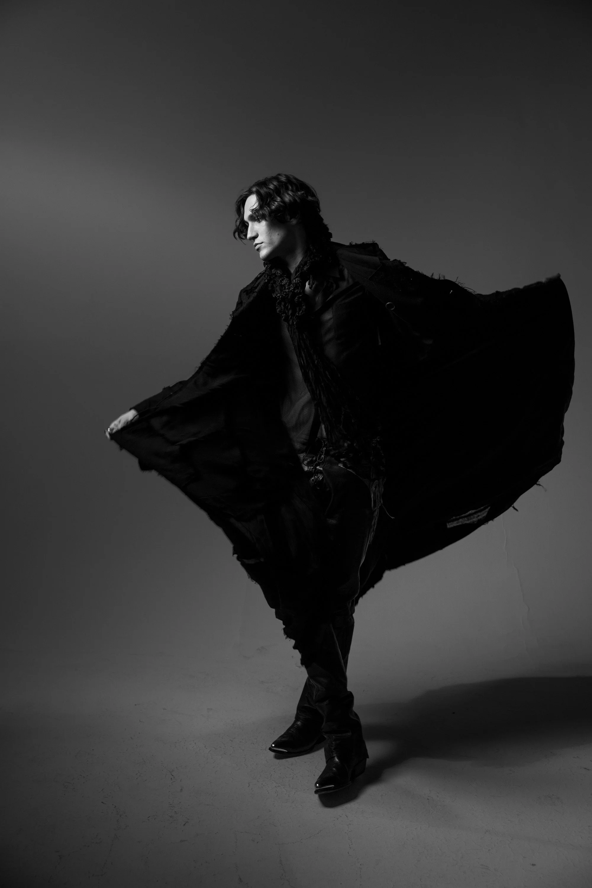 Black and white photo of a person with wavy hair wearing a flowing, dark outfit, mid-dance or pose, against a plain gradient background.