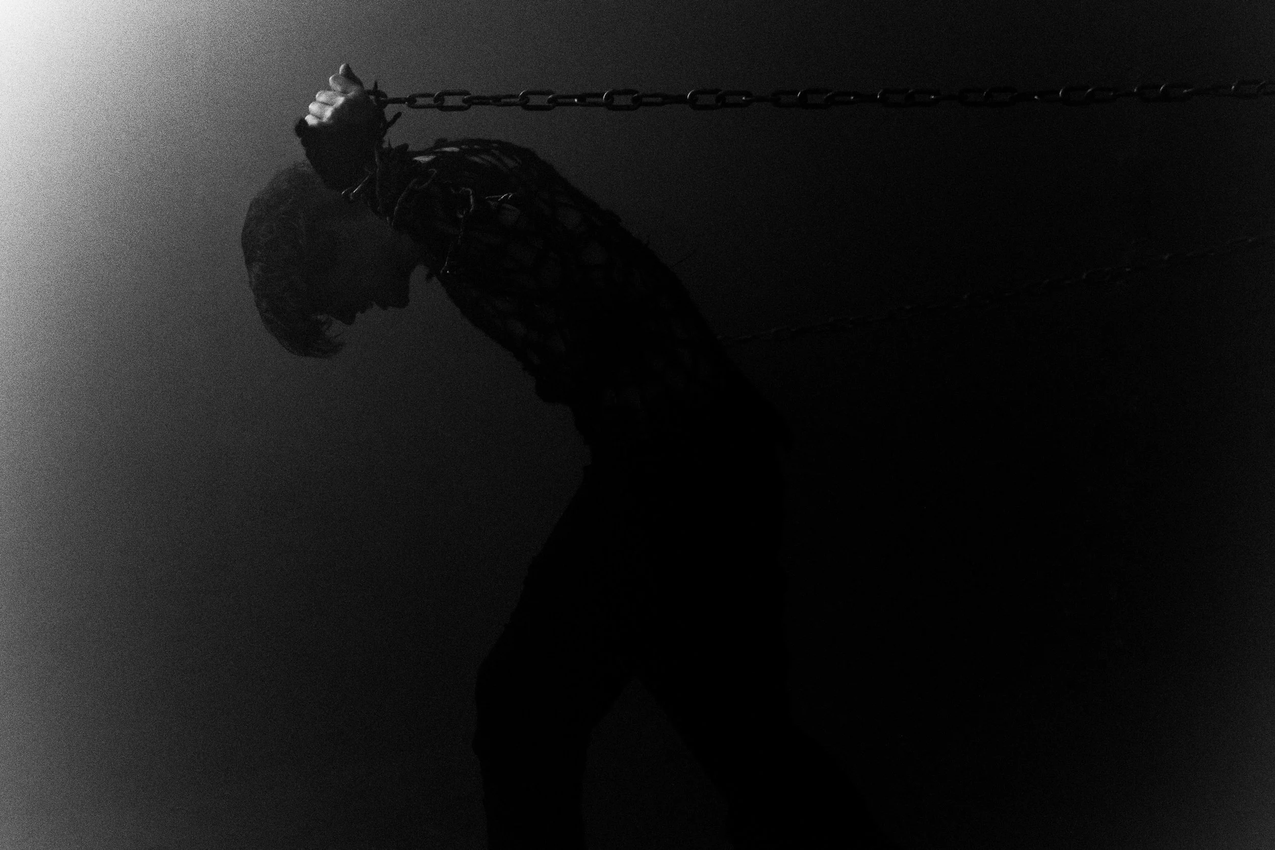 A person holding the chain of a swing set in a dark, minimalistic setting with shadows and no visible background details.