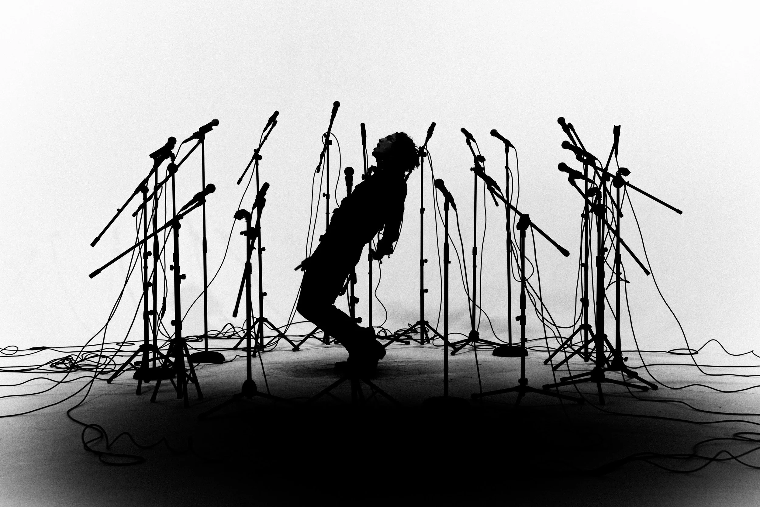 A person standing amidst numerous microphone stands and cords, silhouetted against a plain background.