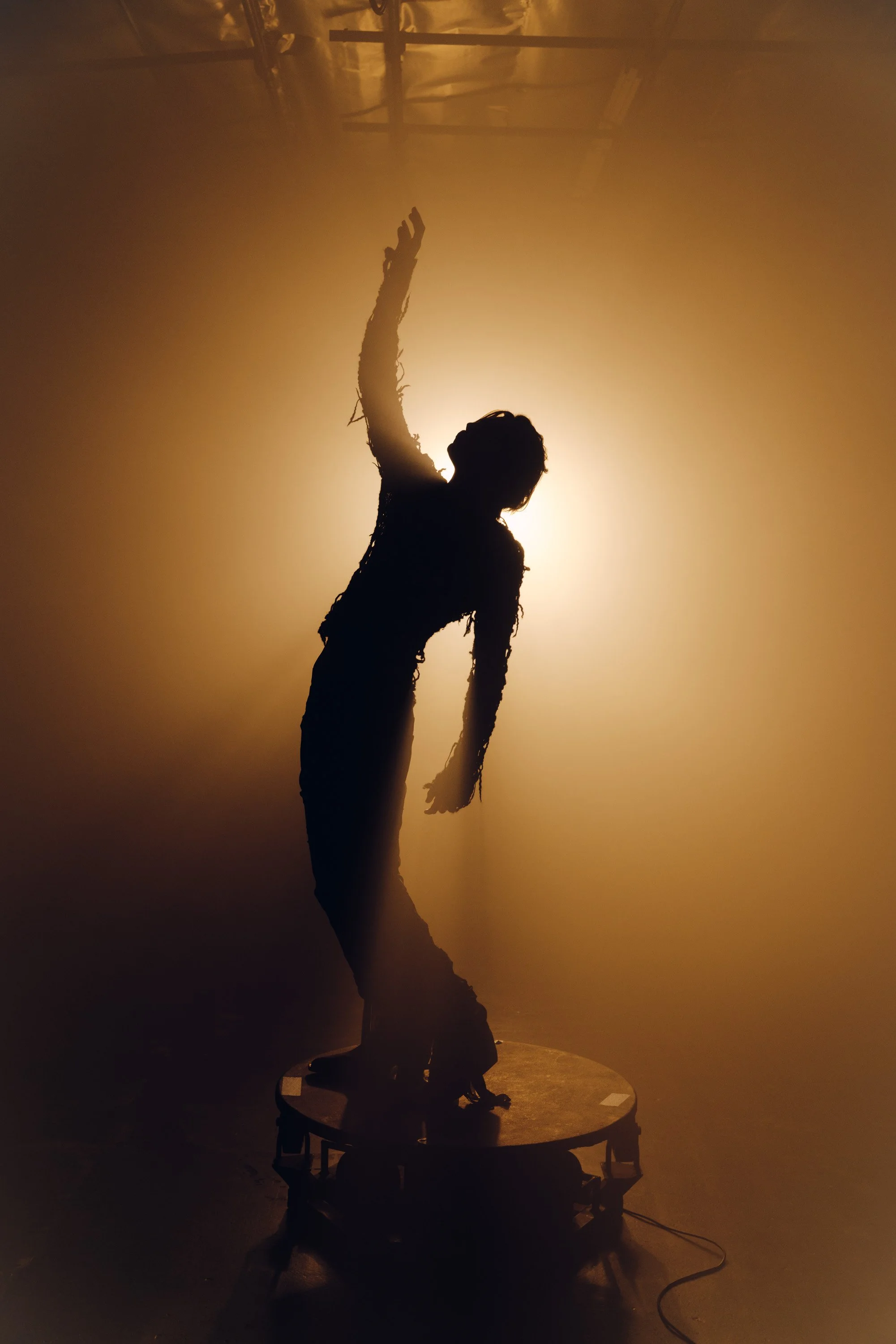 Silhouette of a person standing on a stage, reaching upward, backlit by bright light creating a dramatic shadow effect.