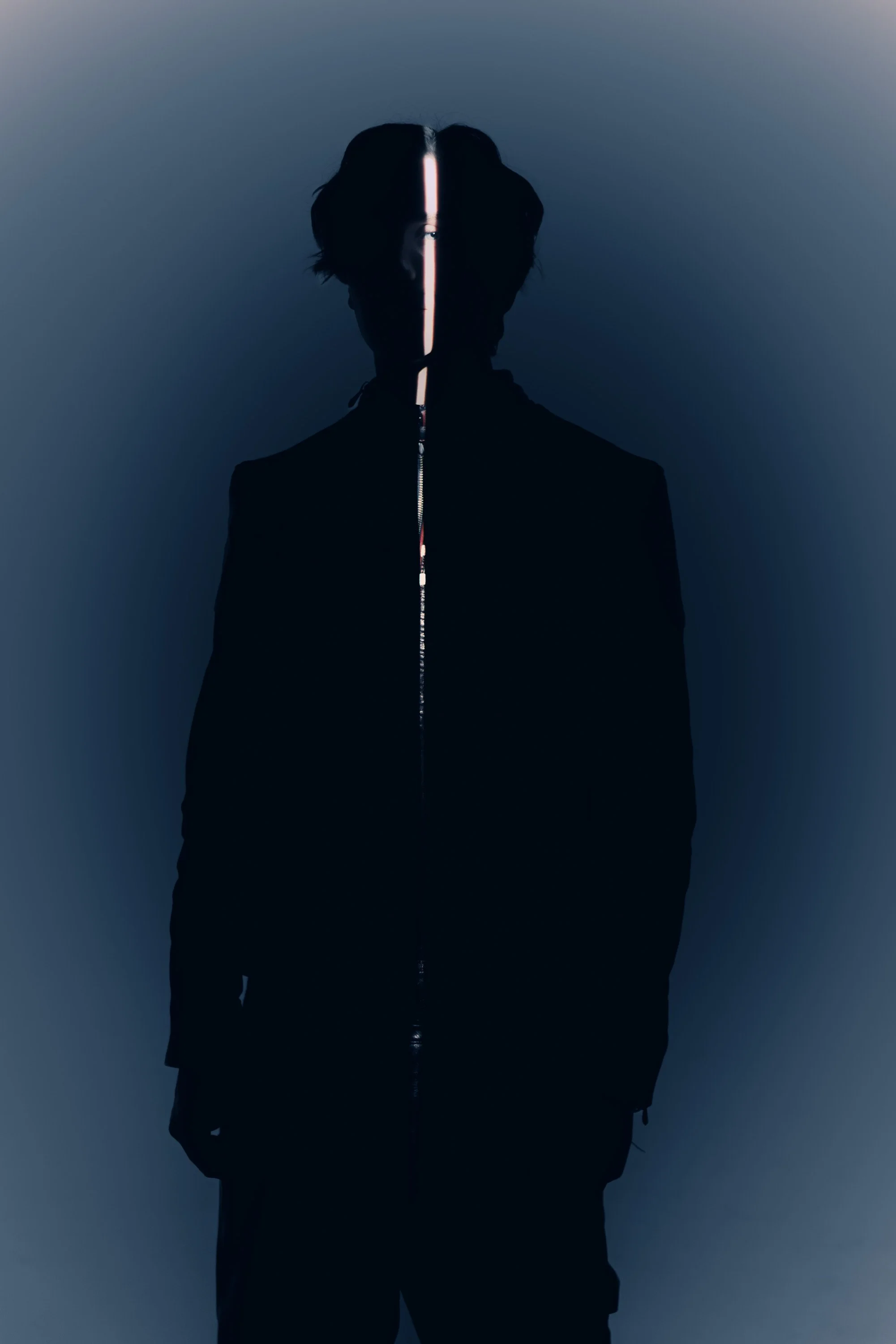 A person in a black jacket standing against a gradient background with a vertical line of light on their face, creating a mysterious, futuristic vibe.