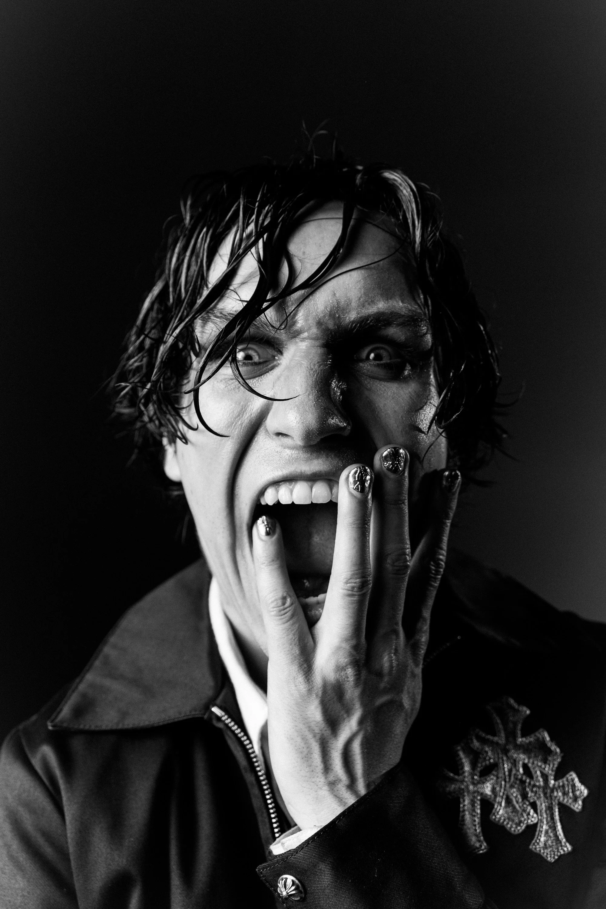 A person with wet, curly hair and intense stare, covering their mouth with their hand, wearing dark clothing with embellishments.