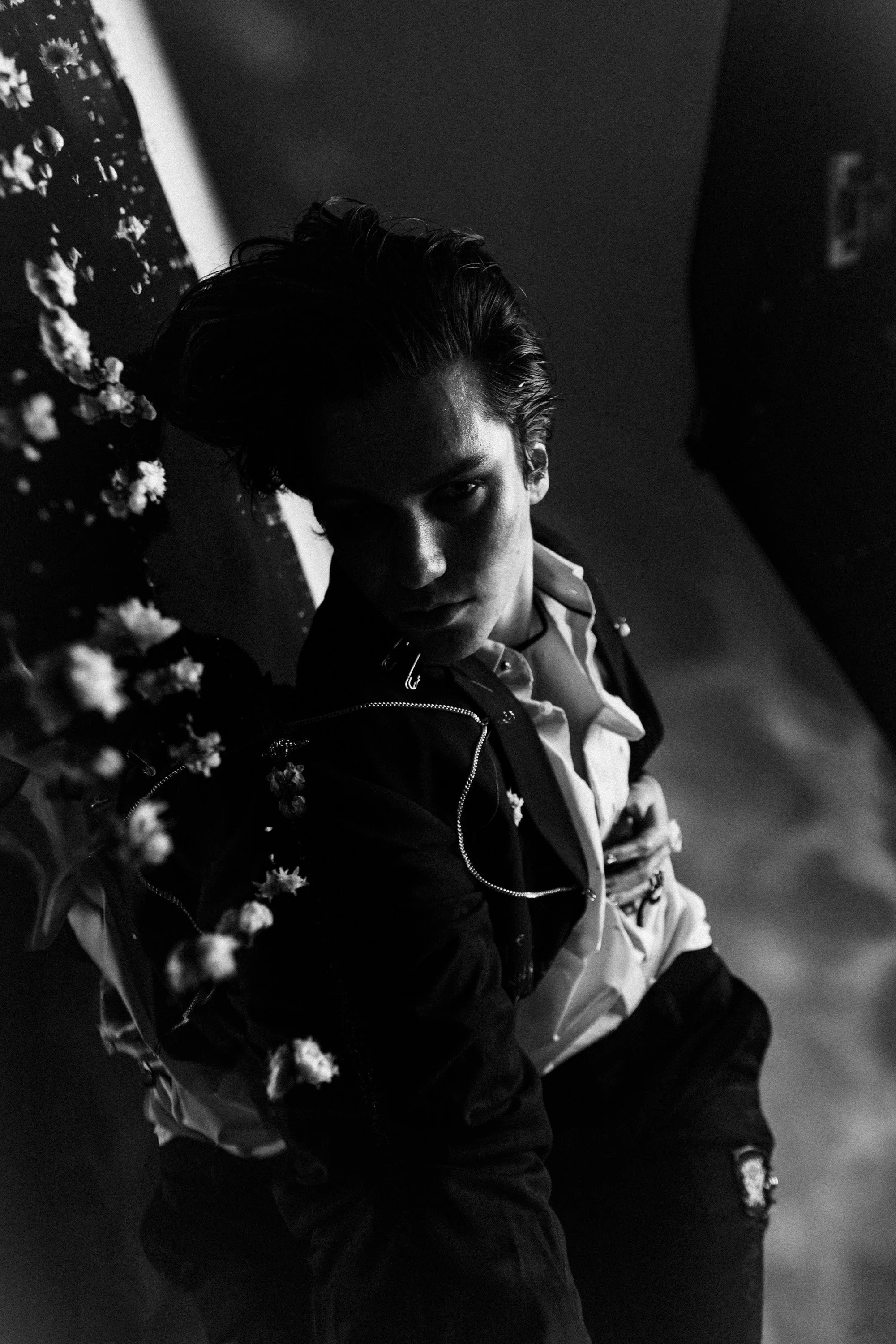 A young man with styled hair wearing a jacket, white shirt, and layered clothing, standing against a dark background with a reflective surface, and flowers visible in the surroundings.