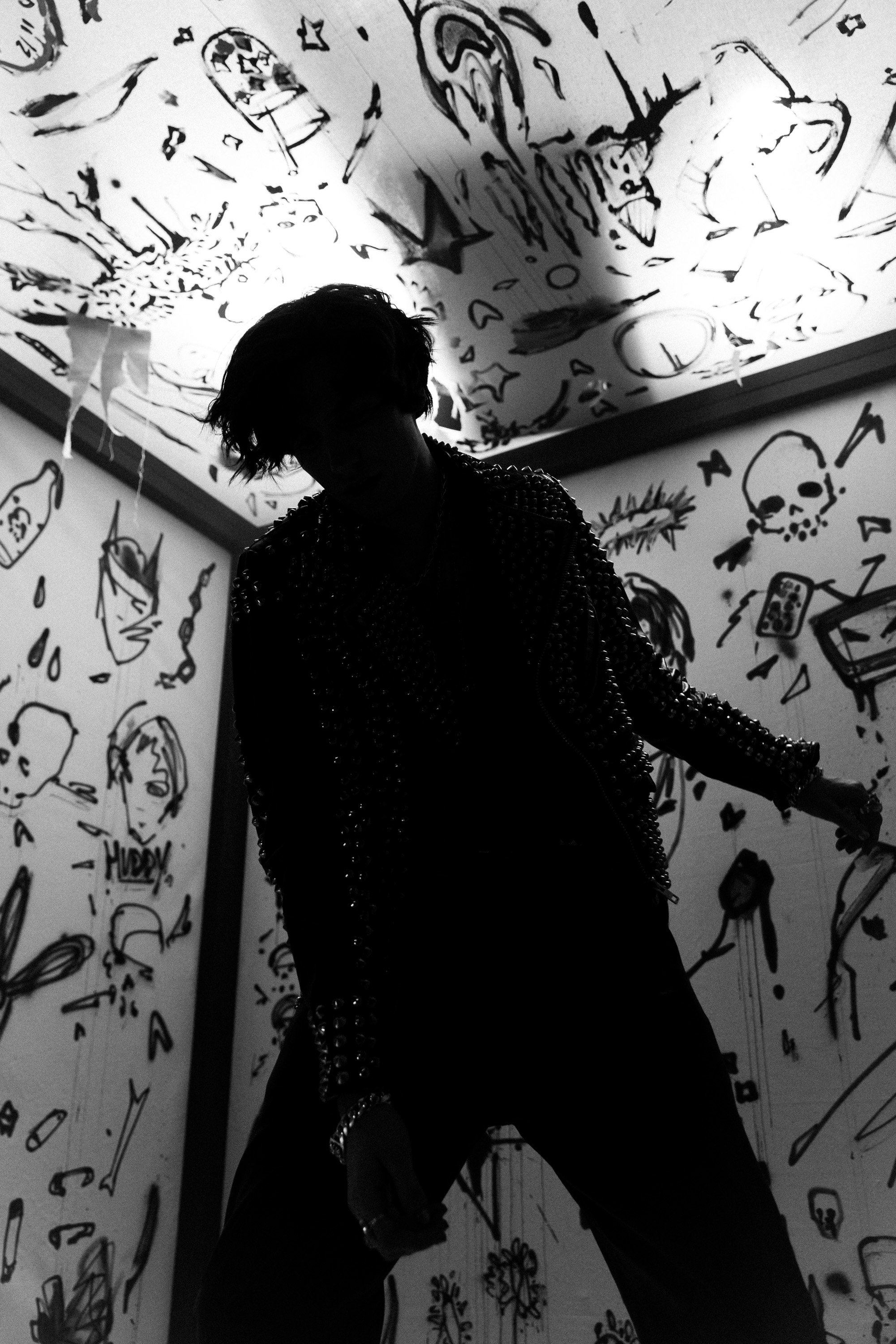 A person with short hair wearing a studded leather jacket and dark pants, standing in a room with walls and ceiling decorated with various black graffiti-style drawings and sketches, including skulls, faces, and abstract designs, all in black and whi