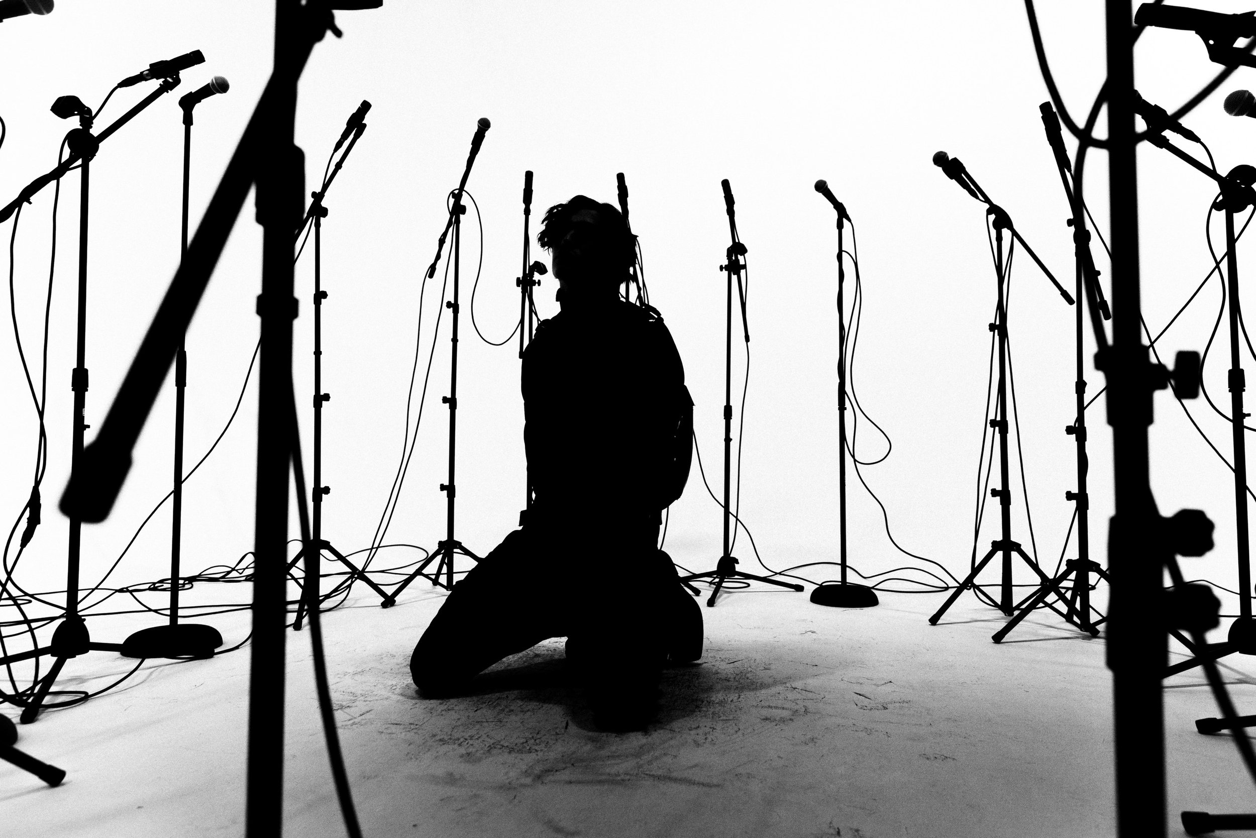 Silhouette of a person kneeling on the ground with numerous microphone stands surrounding them on a white background.