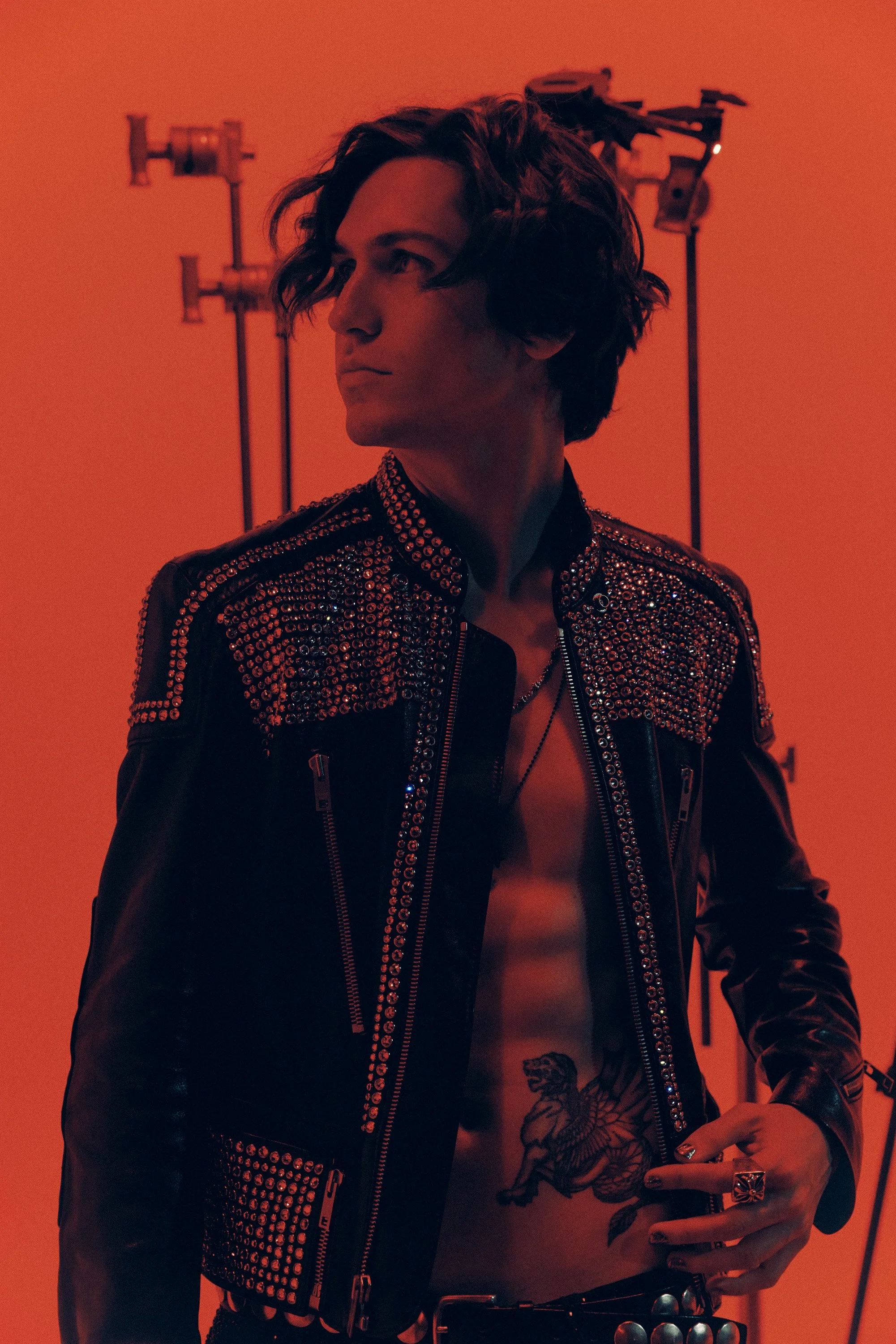 A young man with wavy hair, wearing a black leather jacket with silver studs, standing against an orange background with lighting equipment visible behind him.