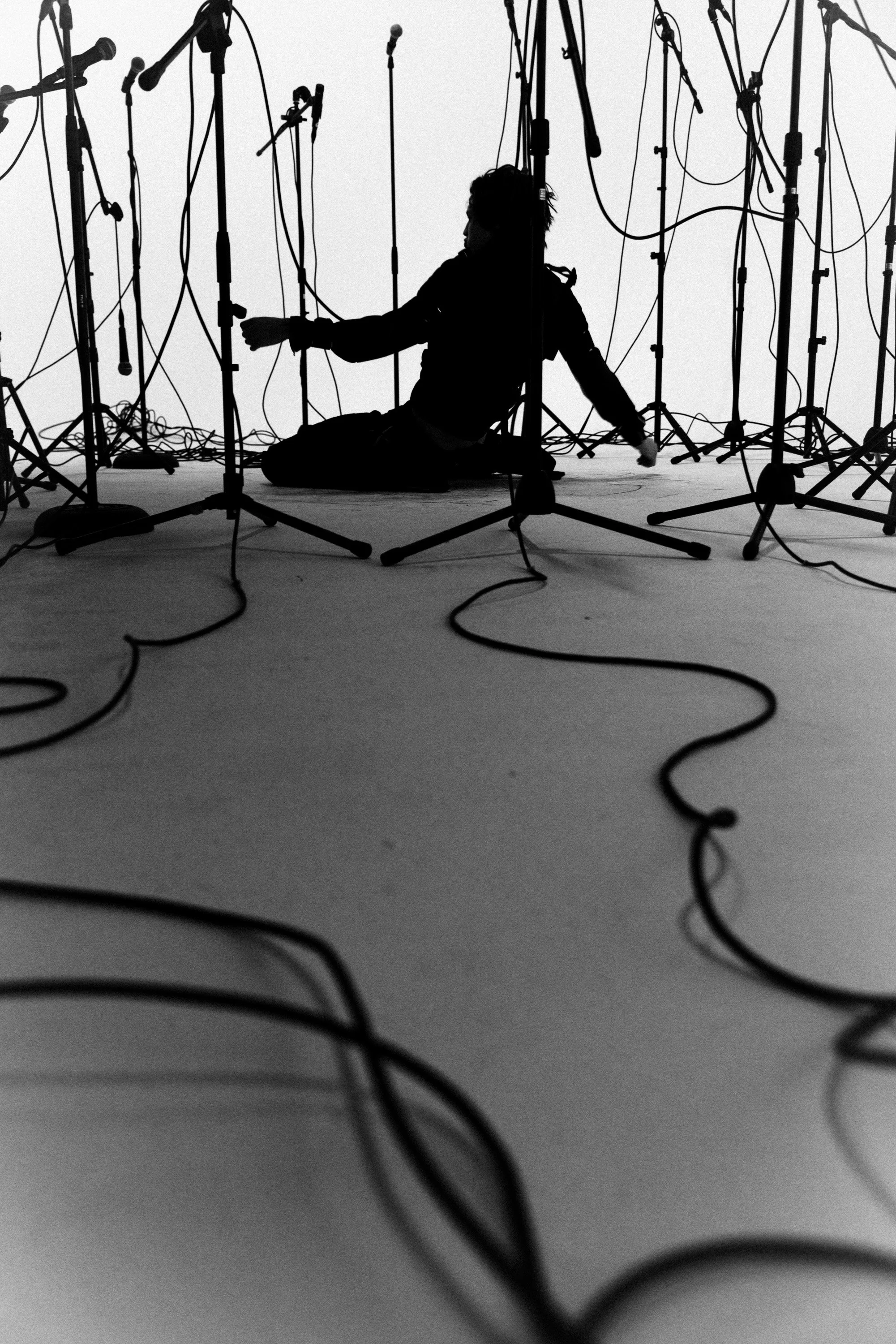 Black silhouette of a person sitting on the floor amidst multiple microphone stands and cables in a recording studio or stage setting.