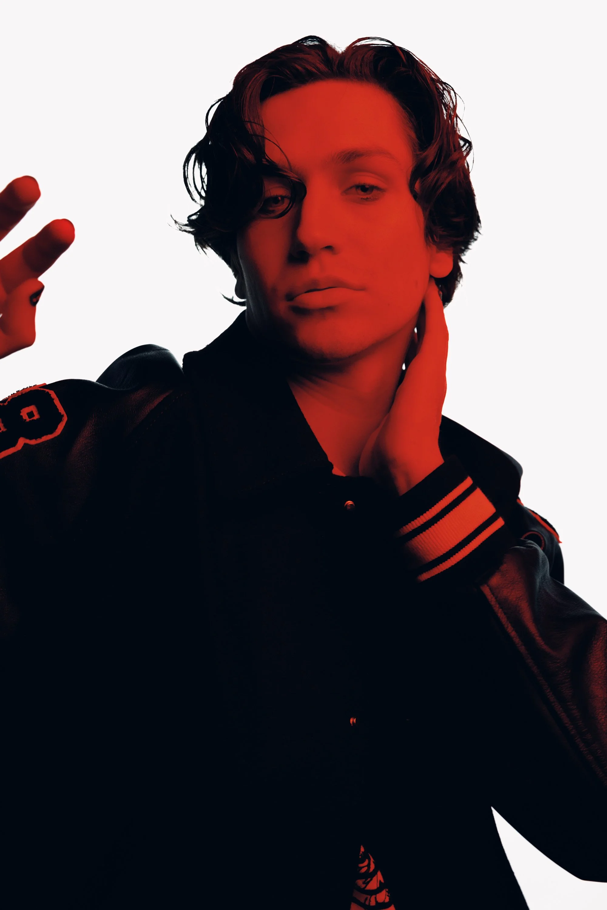 A young man with wavy dark hair, wearing a black jacket with red accents, poses with one hand near his face and the other making a peace sign, set against a white background with red lighting