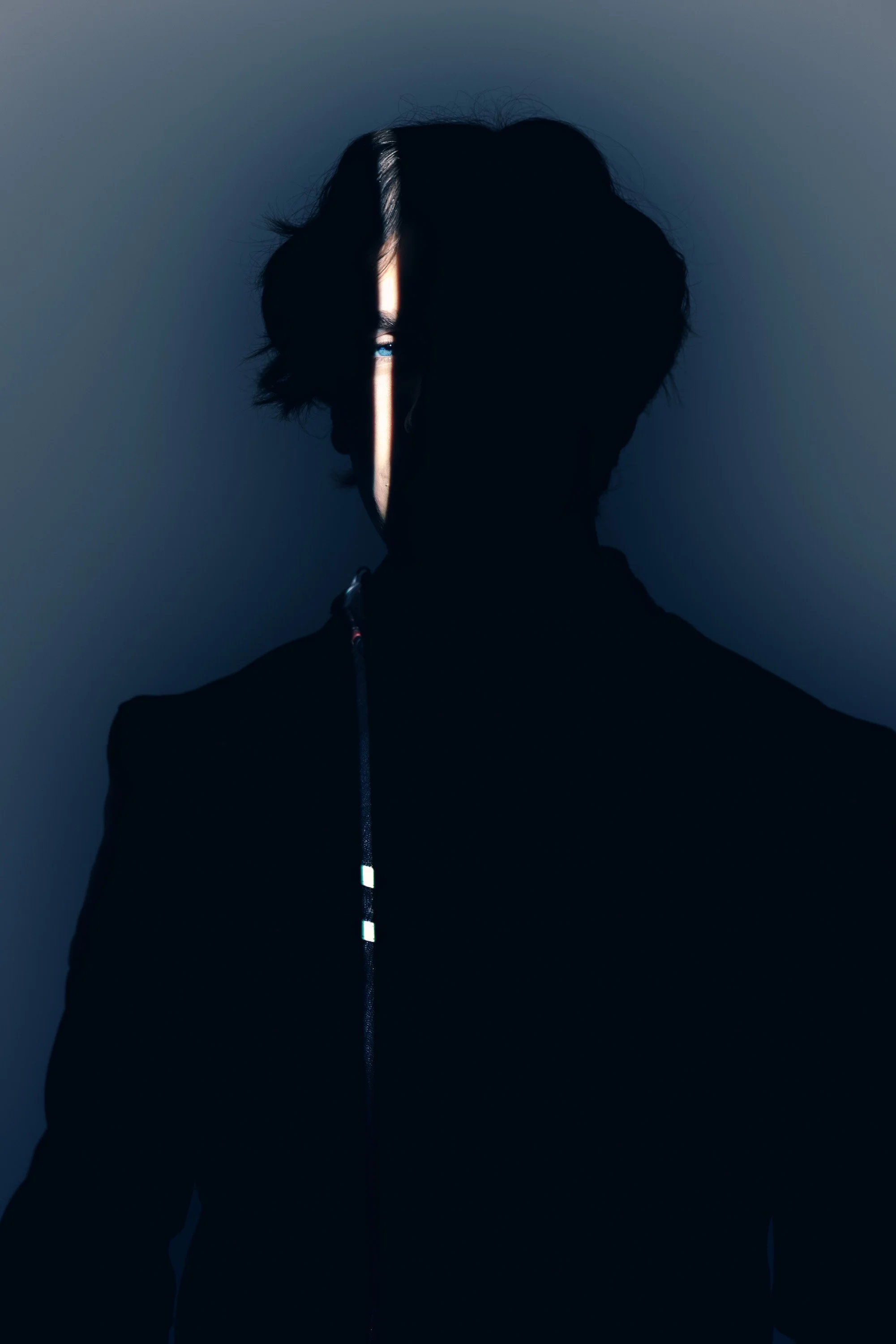 Silhouette of a person with dark, wavy hair and a curtain of light partly illuminating their face against a dark background.