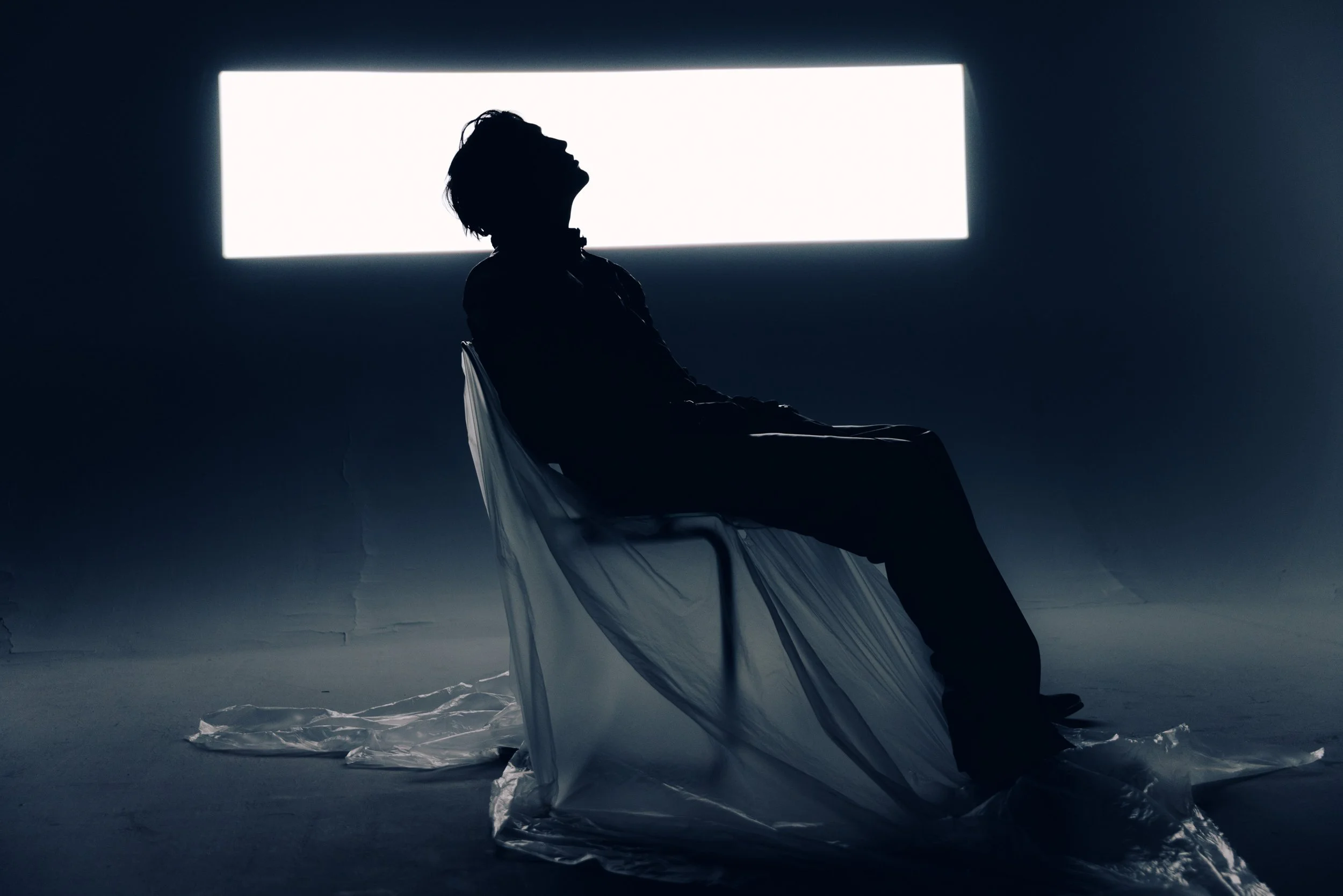 Silhouette of a person sitting on a chair with a large illuminated rectangle behind them, creating a dramatic and abstract scene.