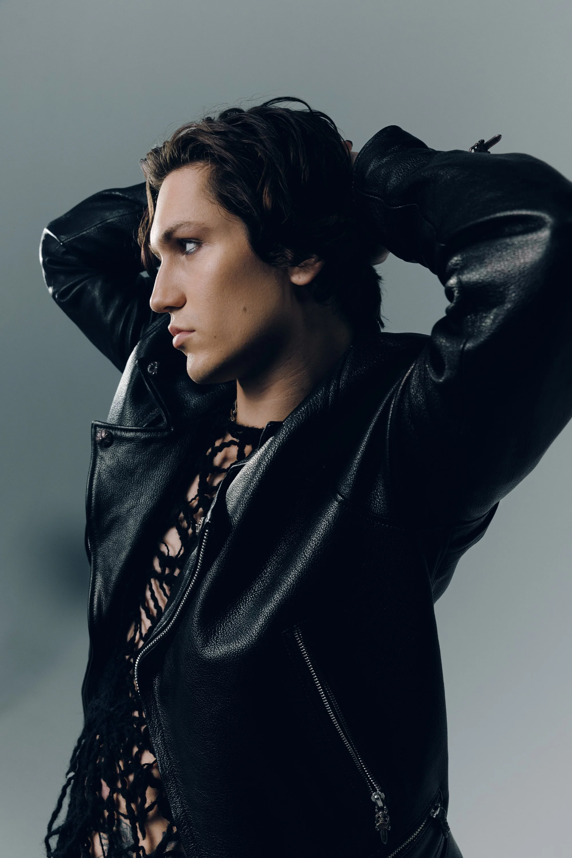 A person with short dark hair wearing a black leather jacket with silver zippers, holding their arms behind their head, against a plain gray background.