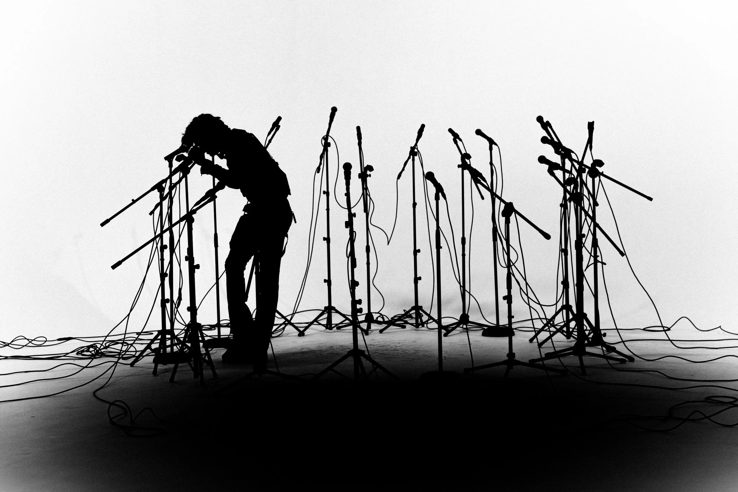 Silhouette of a person working with multiple microphone stands in a studio or stage setting with a plain background.