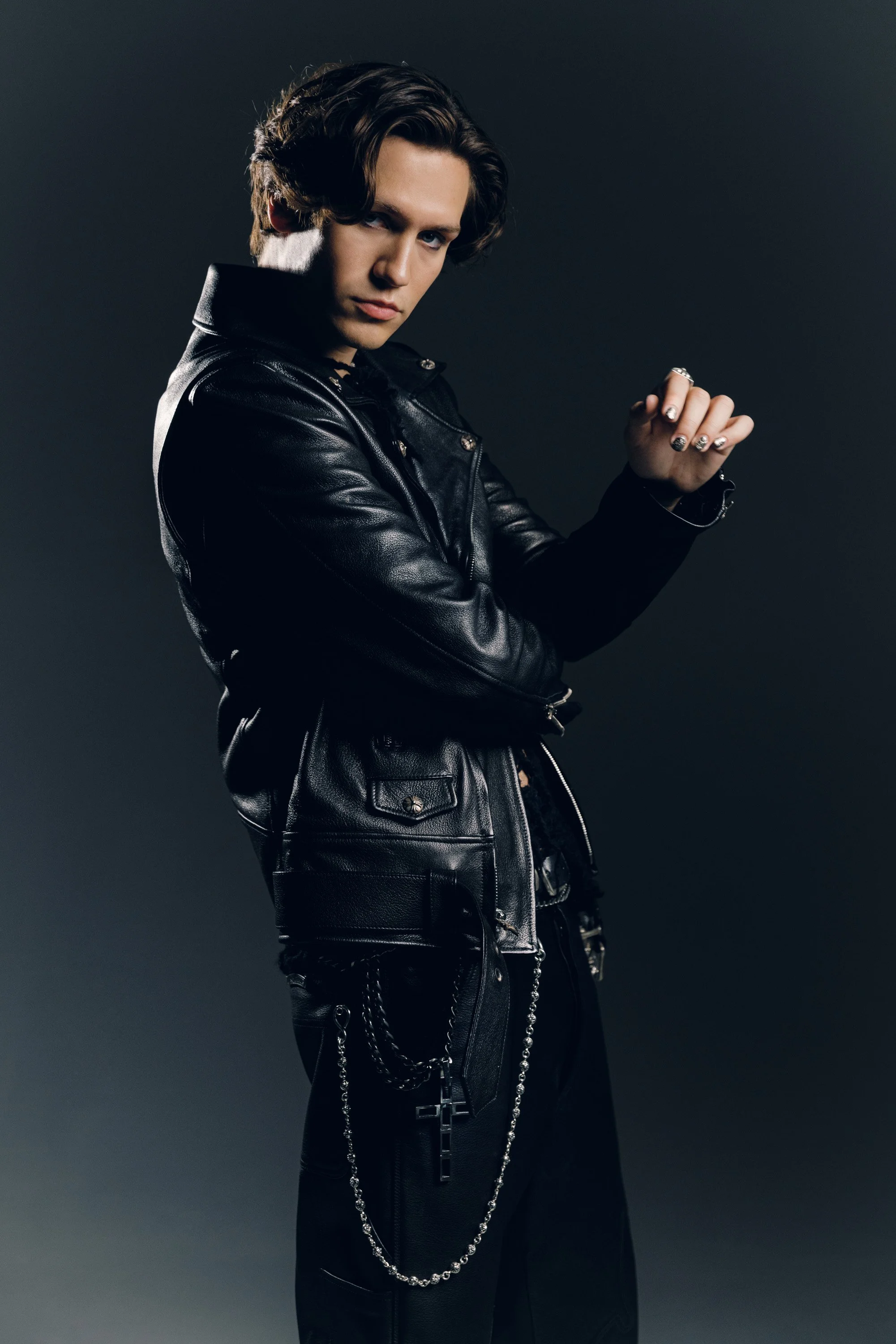 A person with light skin and wavy brown hair styled with volume, wearing a black leather jacket, black pants with chains, and silver rings, poses with one arm across their chest against a dark background.