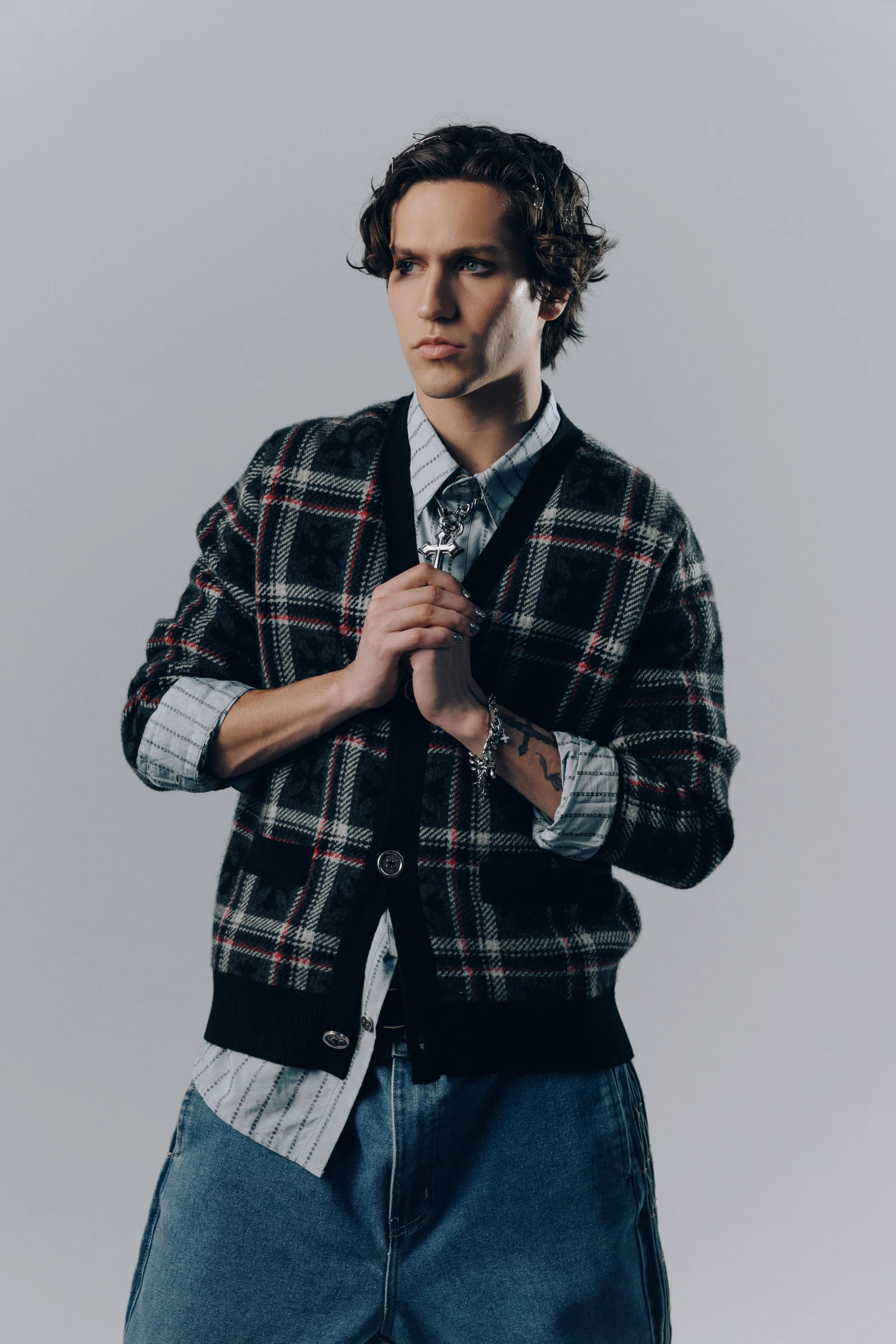 A young man with light skin and wavy dark hair, wearing a plaid cardigan over a striped shirt, with hand clasped together, standing against a plain light gray background.
