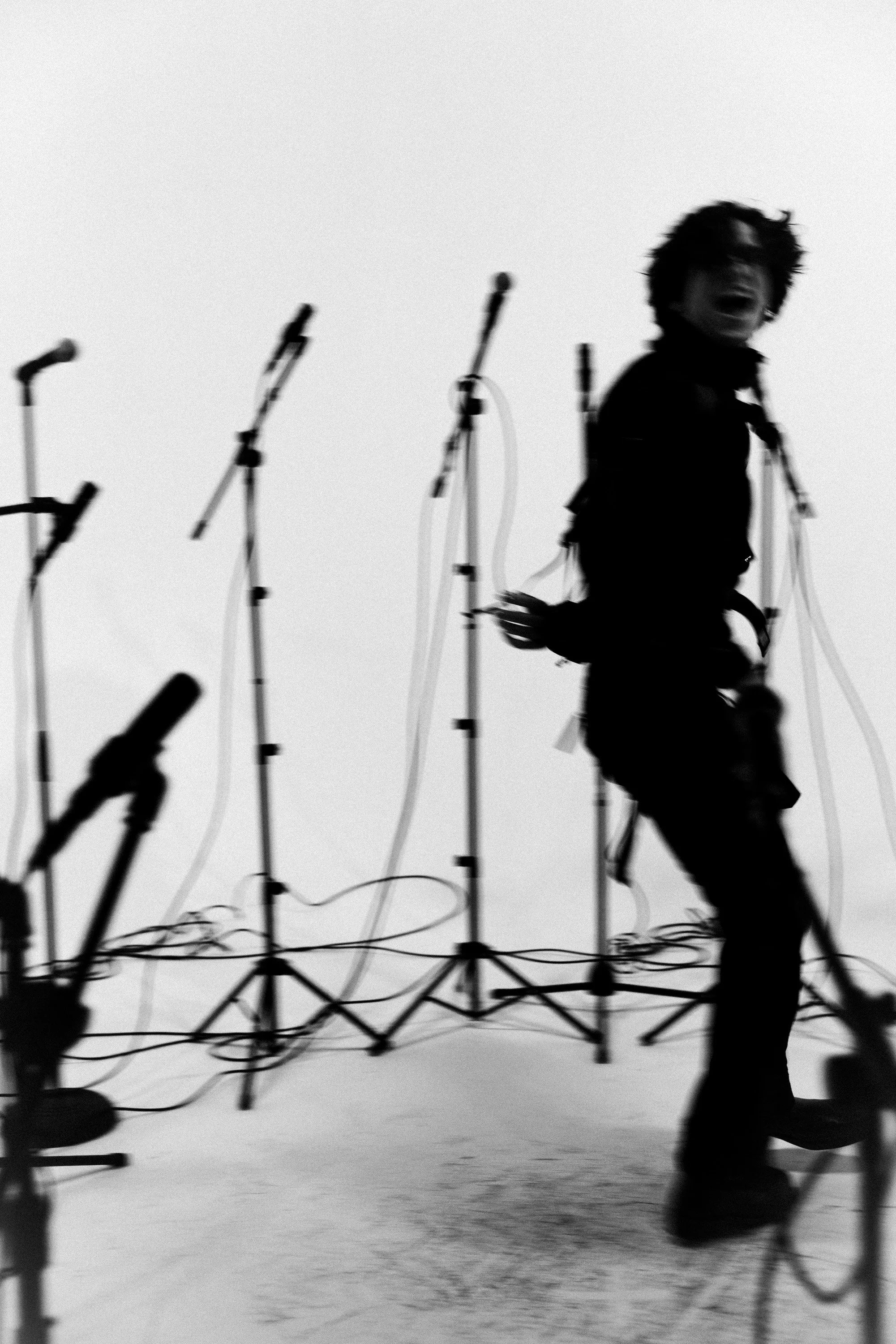 A person running on stage surrounded by multiple microphone stands and cables, with a plain background.