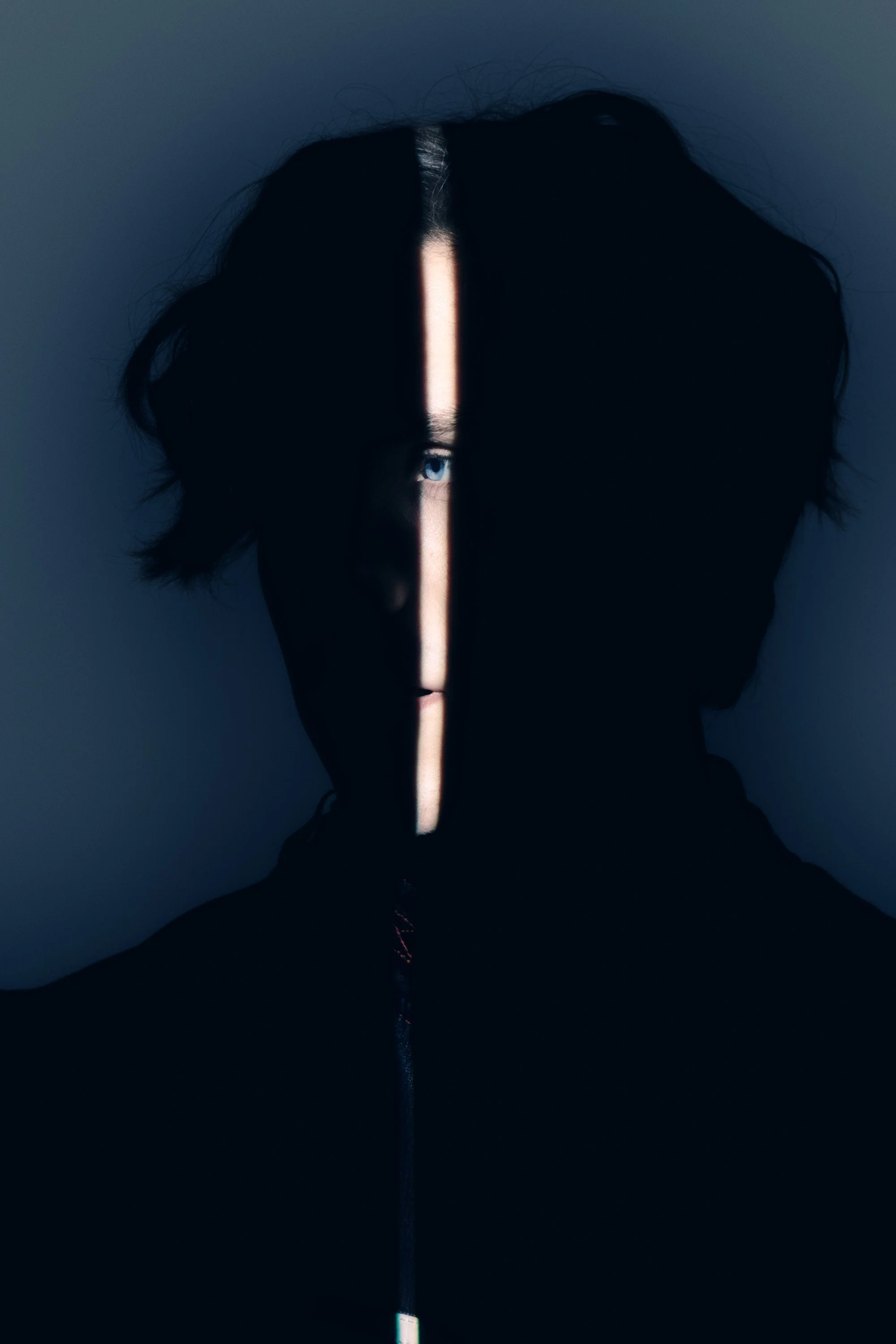 A person with black hair and one visible blue eye is seen partially obscured by darkness, with a vertical streak of light cutting across their face and revealing one eye.