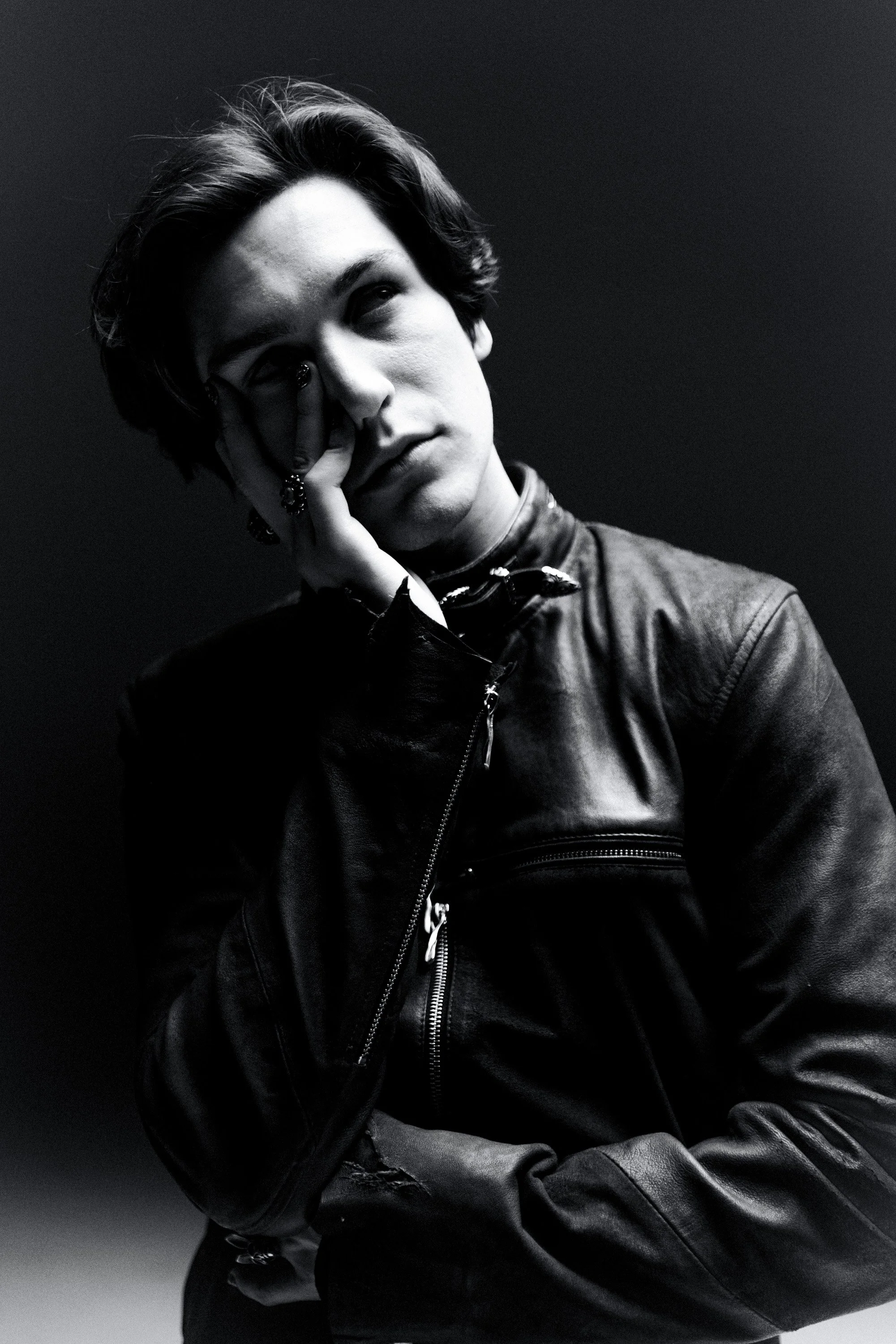 Black and white photograph of a young man with medium-length hair, wearing a leather jacket, sitting against a dark background. He has a contemplative expression, with one hand resting on his face and eyes looking upward.