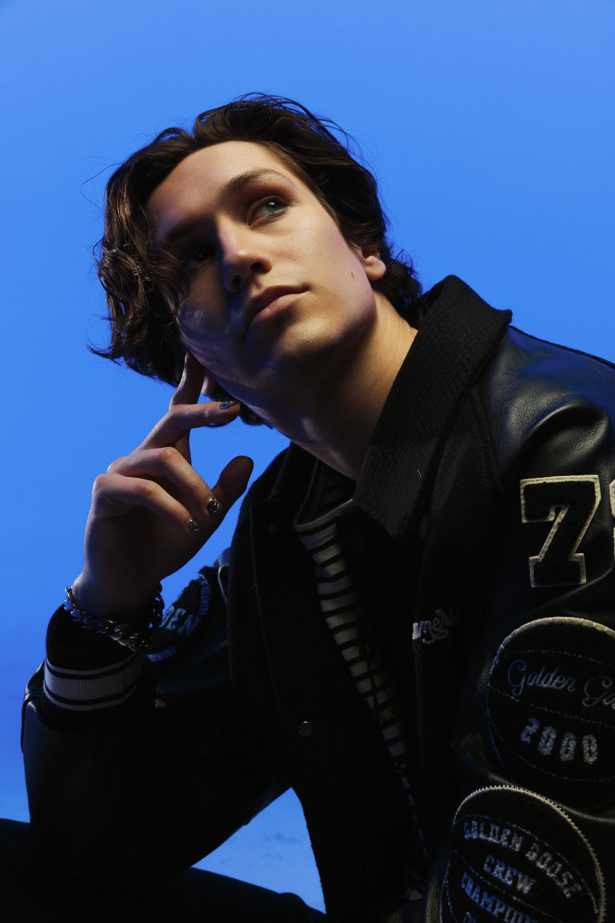 Close-up of a young man with long, wavy brown hair and light skin, posing with his hand touching his face against a blue background. He is wearing a black jacket with patches and a striped shirt underneath.